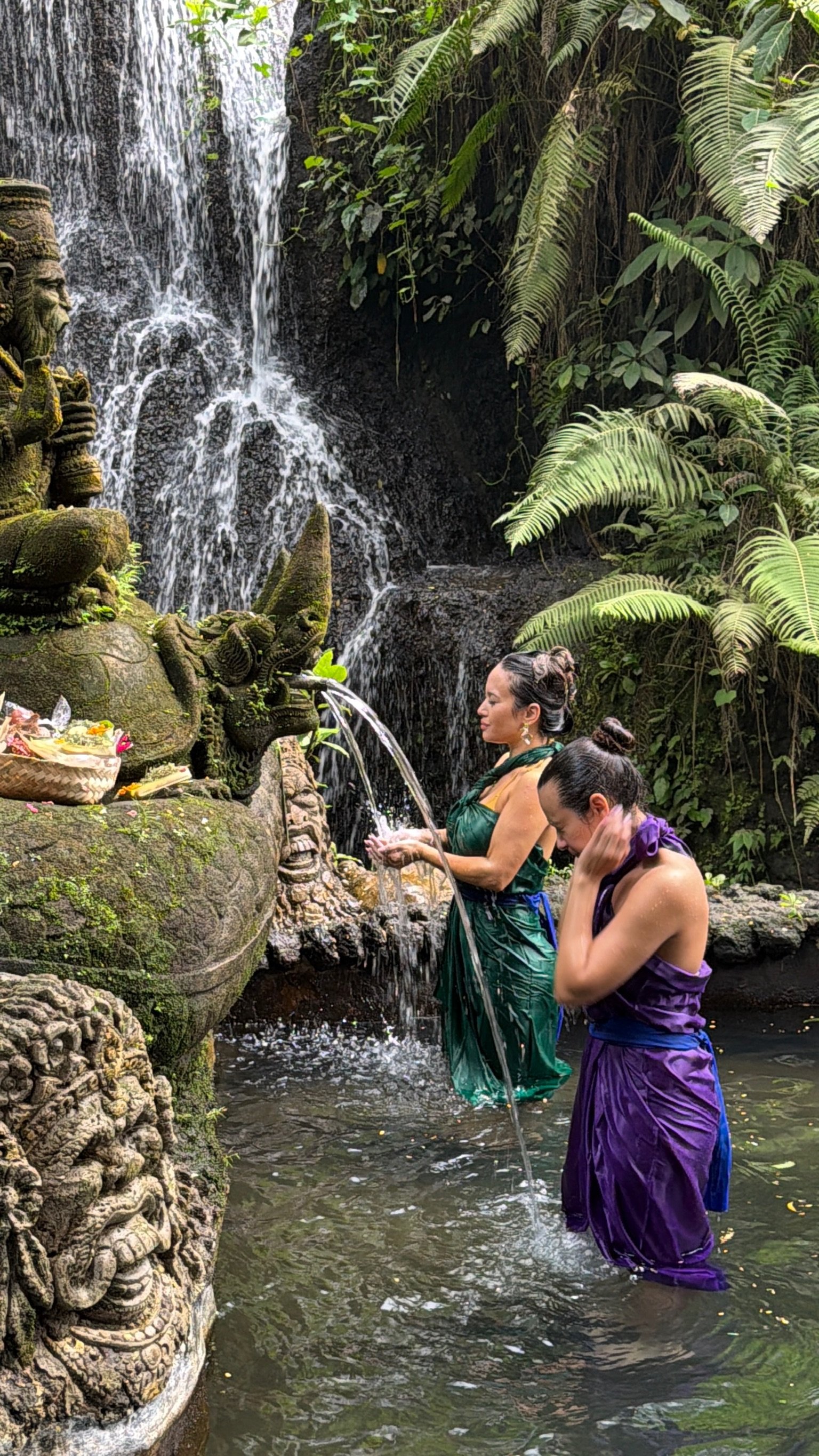 Two women in colorful traditional dresses standing in a water pool at a waterfall, participating in a spiritual water ritual at a stone shrine surrounded by lush green foliage.