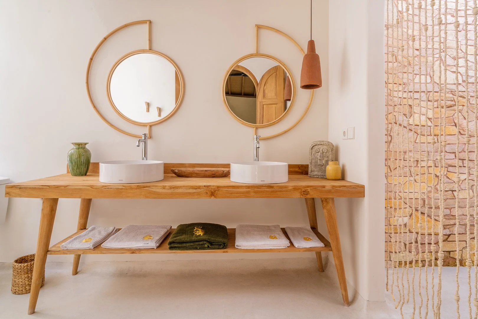 Bathroom with two round mirrors, wooden vanity with two white vessel sinks, decorative items including a green vase, wooden bowl, stone face sculpture, yellow vase, and towels beneath the vanity, and a beaded partition on the right.