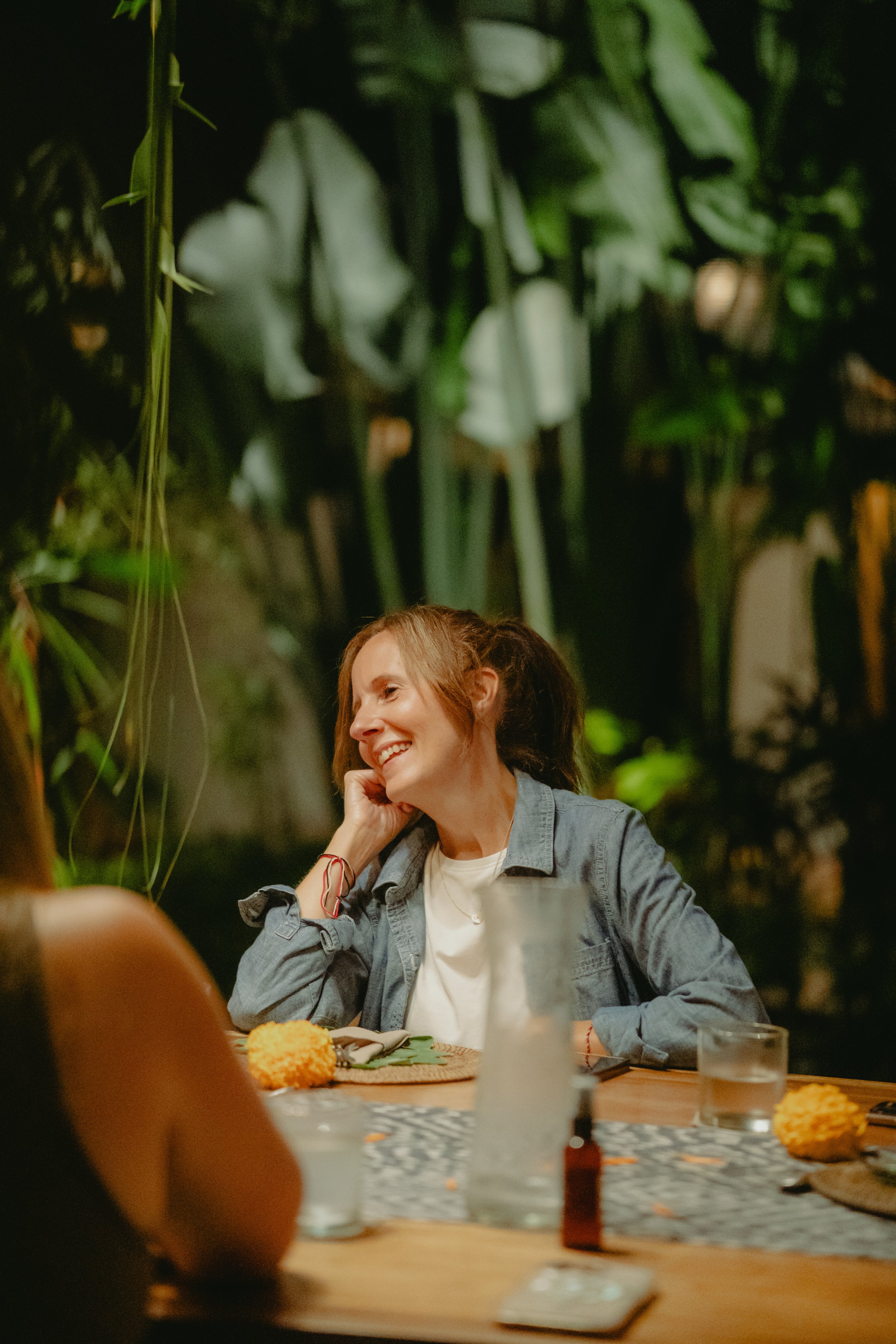 A woman with red hair, smiling and resting her hand on her face, sitting at a table with food and drinks in a dimly lit restaurant or bar with tropical plants in the background.
