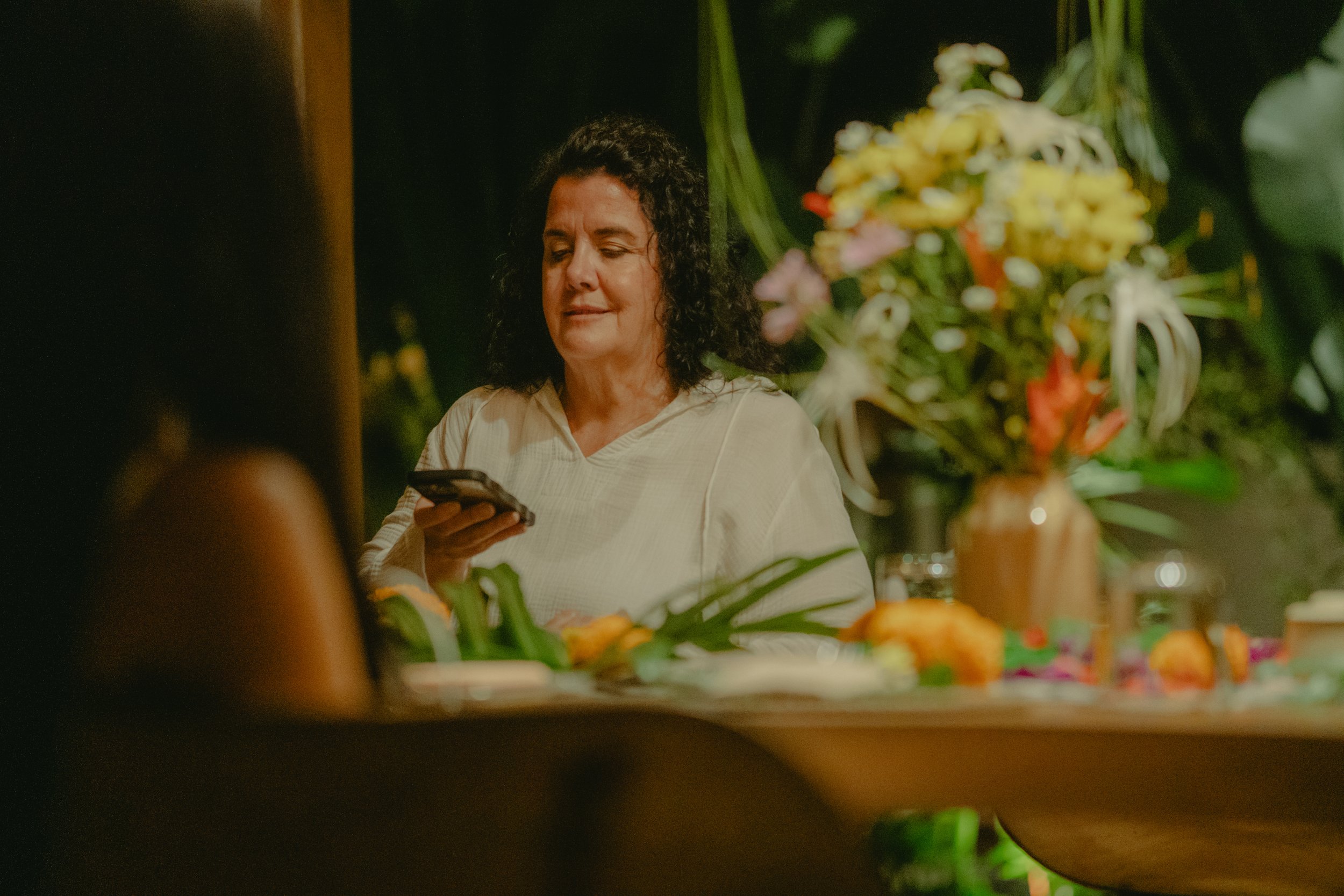 A woman with curly dark hair looking at her phone while standing near a table with flowers and food at a gathering, with a dark background.