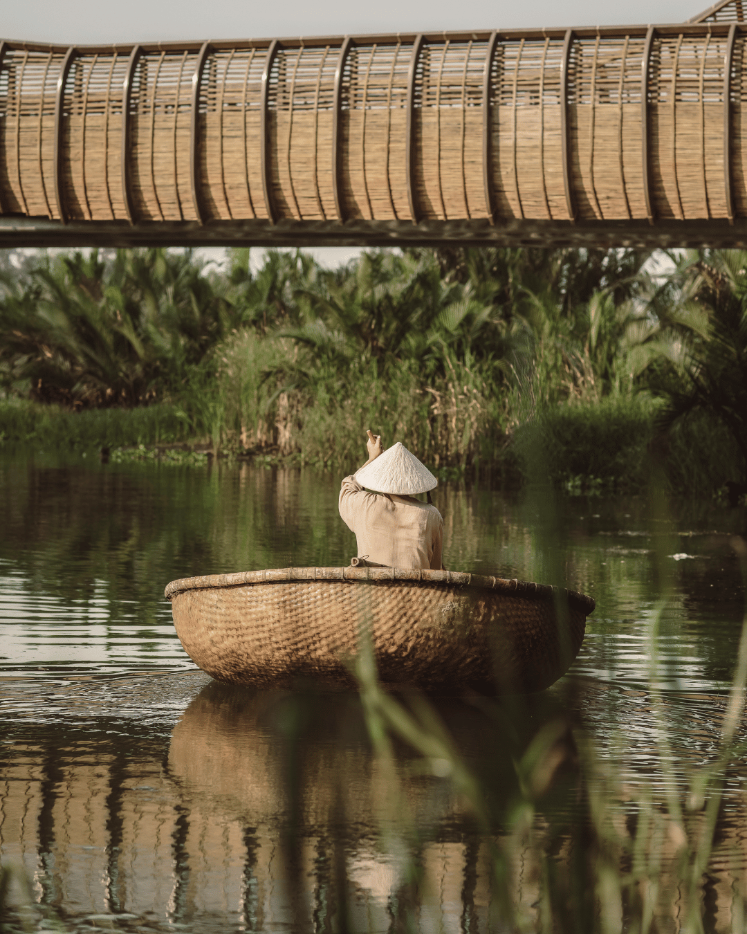Person in traditional attire and conical hat sailing a round basket boat on a calm river surrounded by lush green vegetation, seen from under a wooden bridge. | Awakened Soul - Jessika Harrison 