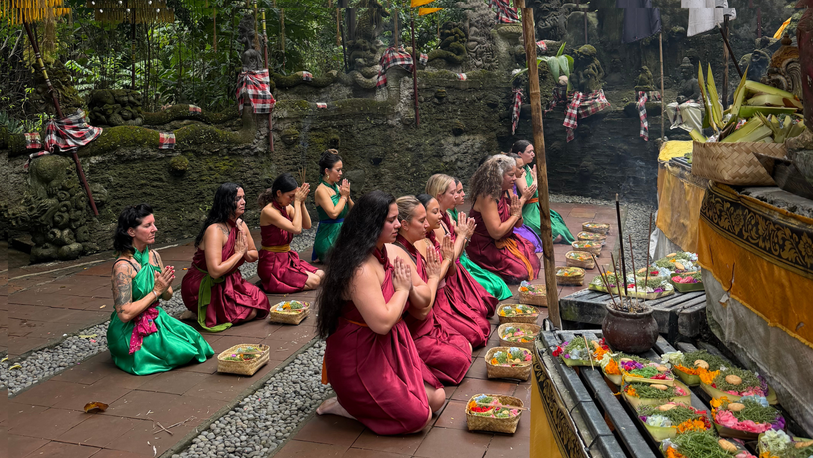 A group of women dressed in traditional Balinese attire kneeling and praying during a religious ceremony outdoors. They are surrounded by offerings of flowers and food in baskets, with stone statues and lush greenery in the background.