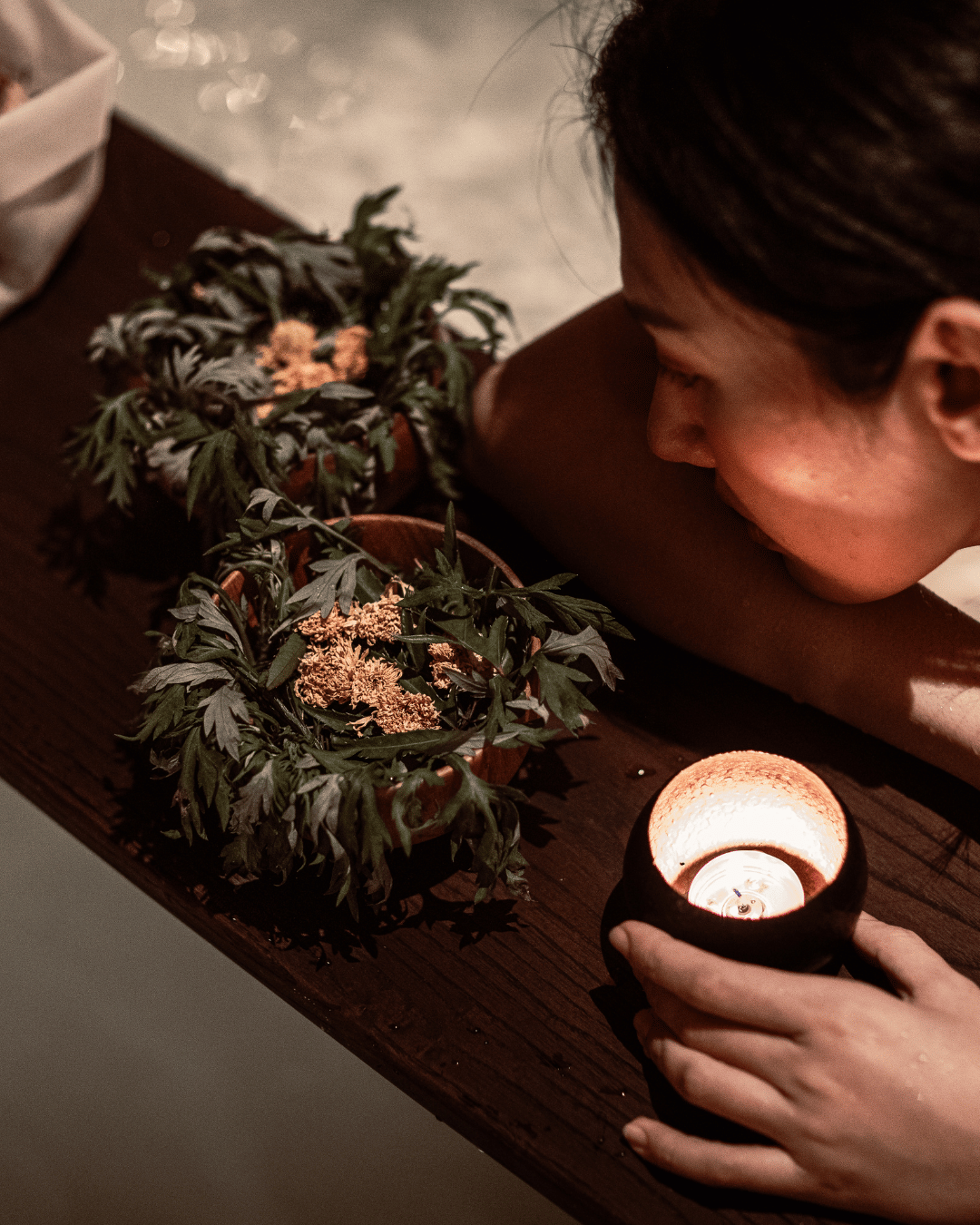 A woman lying on a wooden table with her face close to a table centerpiece made of green leaves and pink flowers, and holding a lit candle in a black holder. | Awakened Soul - Jessika Harrison 