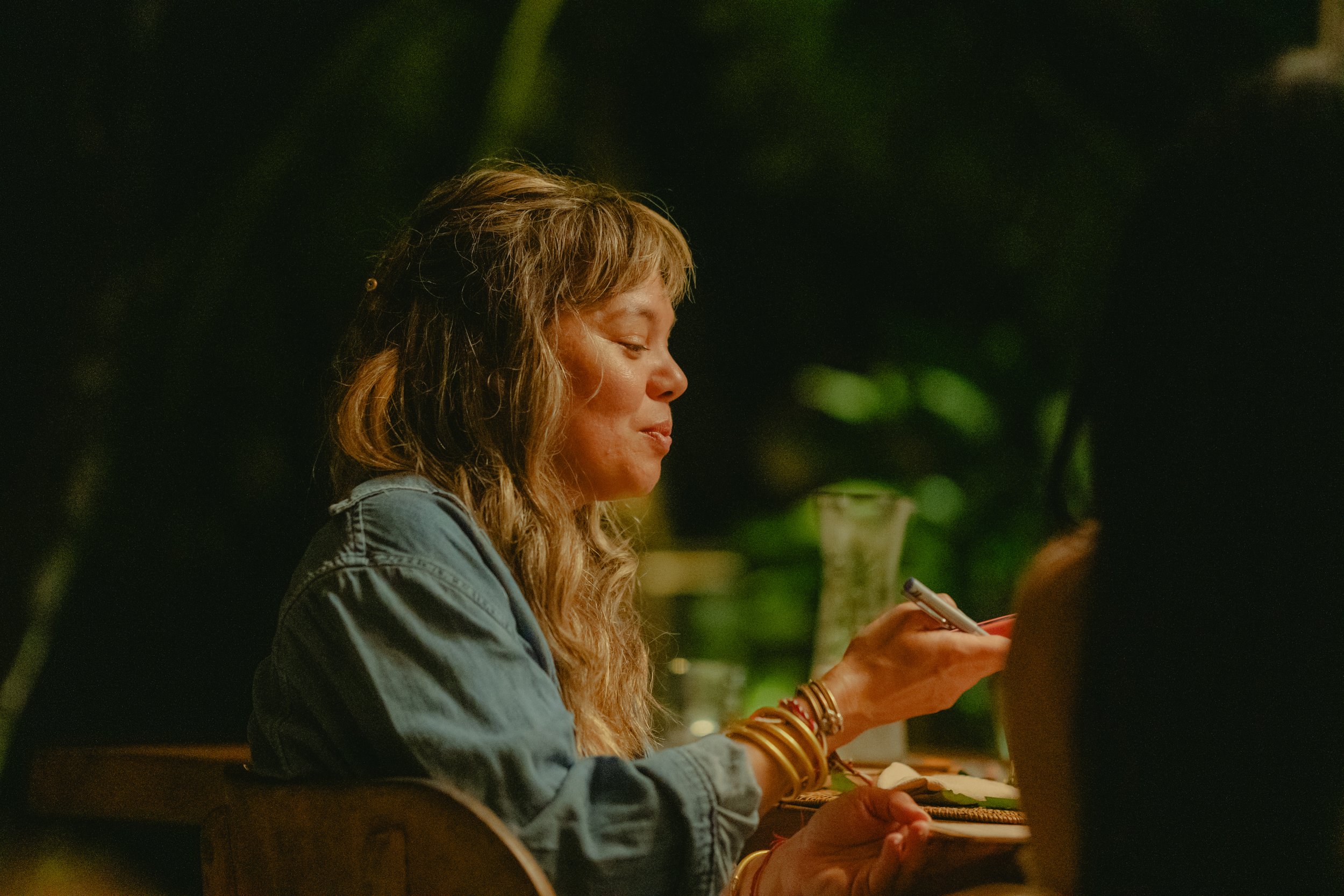 A woman with light brown hair and wearing bracelets on her left wrist, sitting at a table and looking at her phone with a slight smile, in a dimly lit setting with blurred greenery in the background.