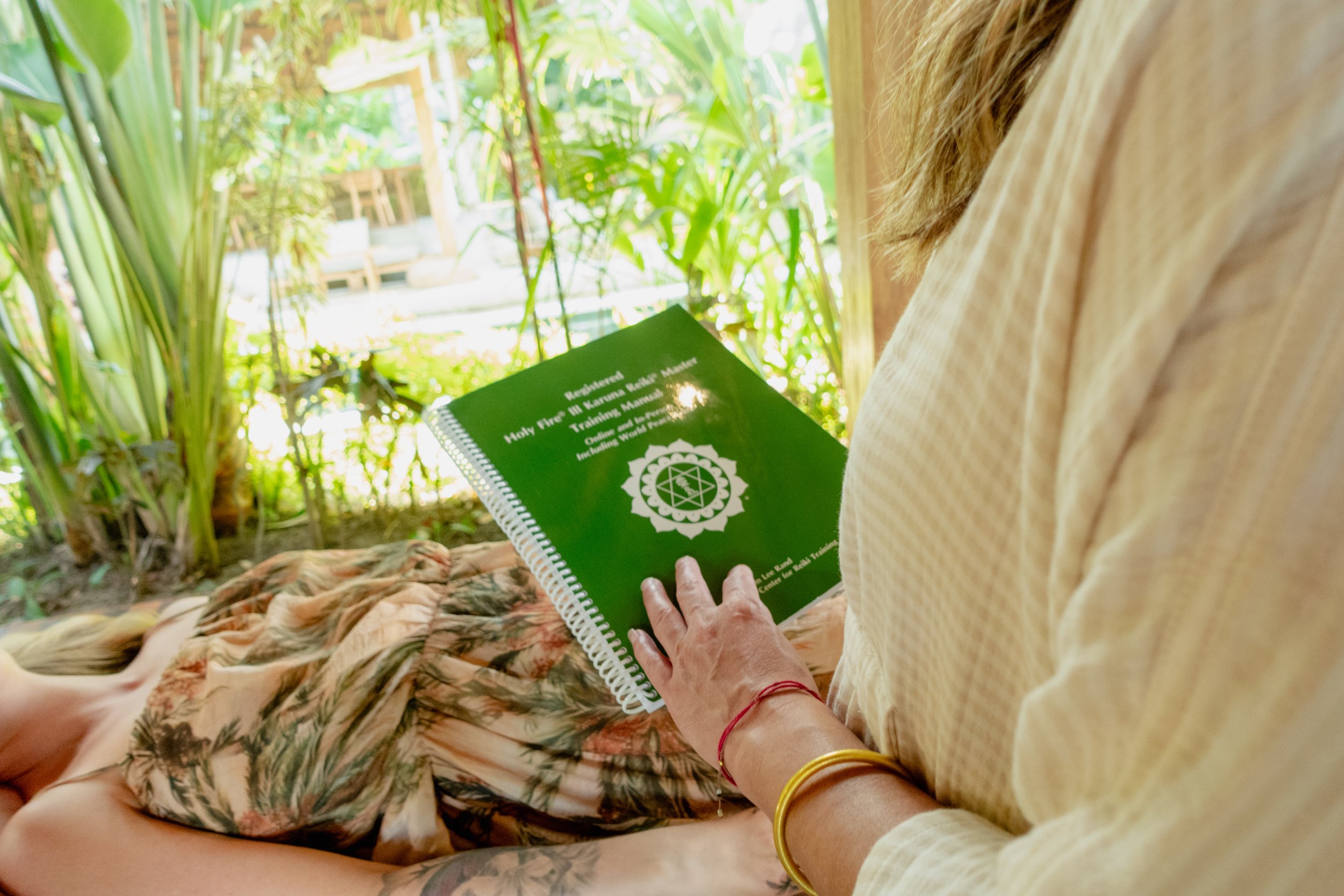 A person holding a green booklet titled 'Holy Fire in Karuna Reiki Master' while sitting outdoors on a floral blanket surrounded by lush tropical plants.