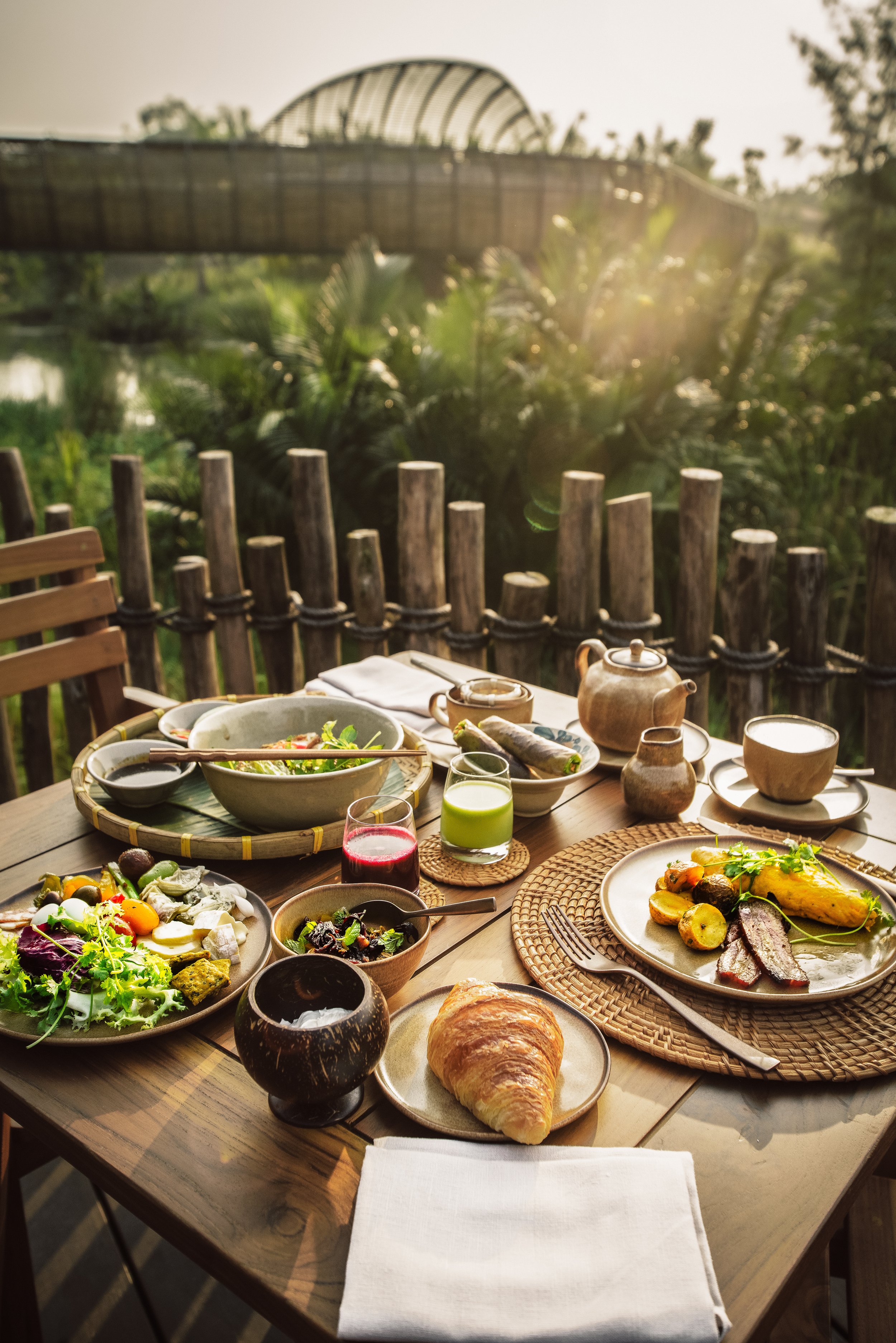 A table set for breakfast outdoors with various dishes, drinks, and utensils, overlooking a lush garden and a river at sunrise. | Awakened Soul - Jessika Harrison 