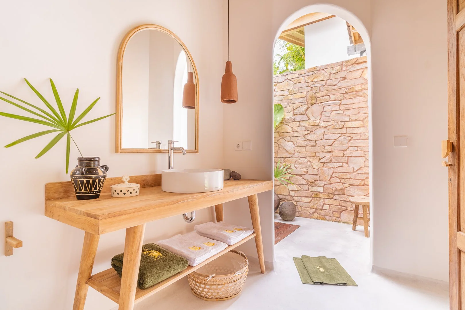 Bathroom with a wooden vanity, round white sink, mirror, hanging pendant lights, potted plant, and outdoor stone patio with stool and potted plants