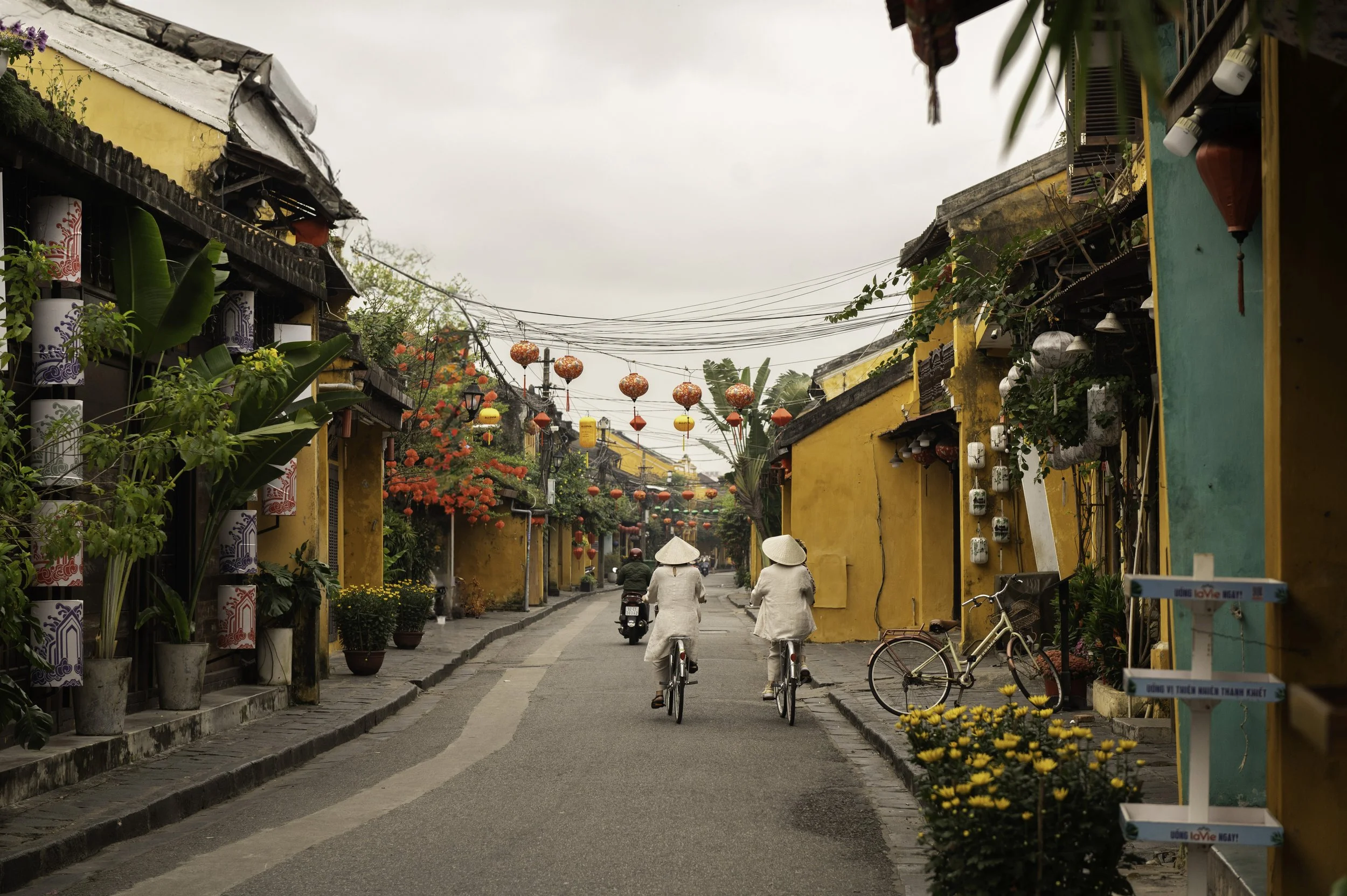 A street scene in a traditional Vietnamese town with colorful lanterns hanging overhead. Two women riding bicycles, walking on the sidewalk, yellow buildings with plants and flowers lining the street, and a person riding a motorbike in the background
