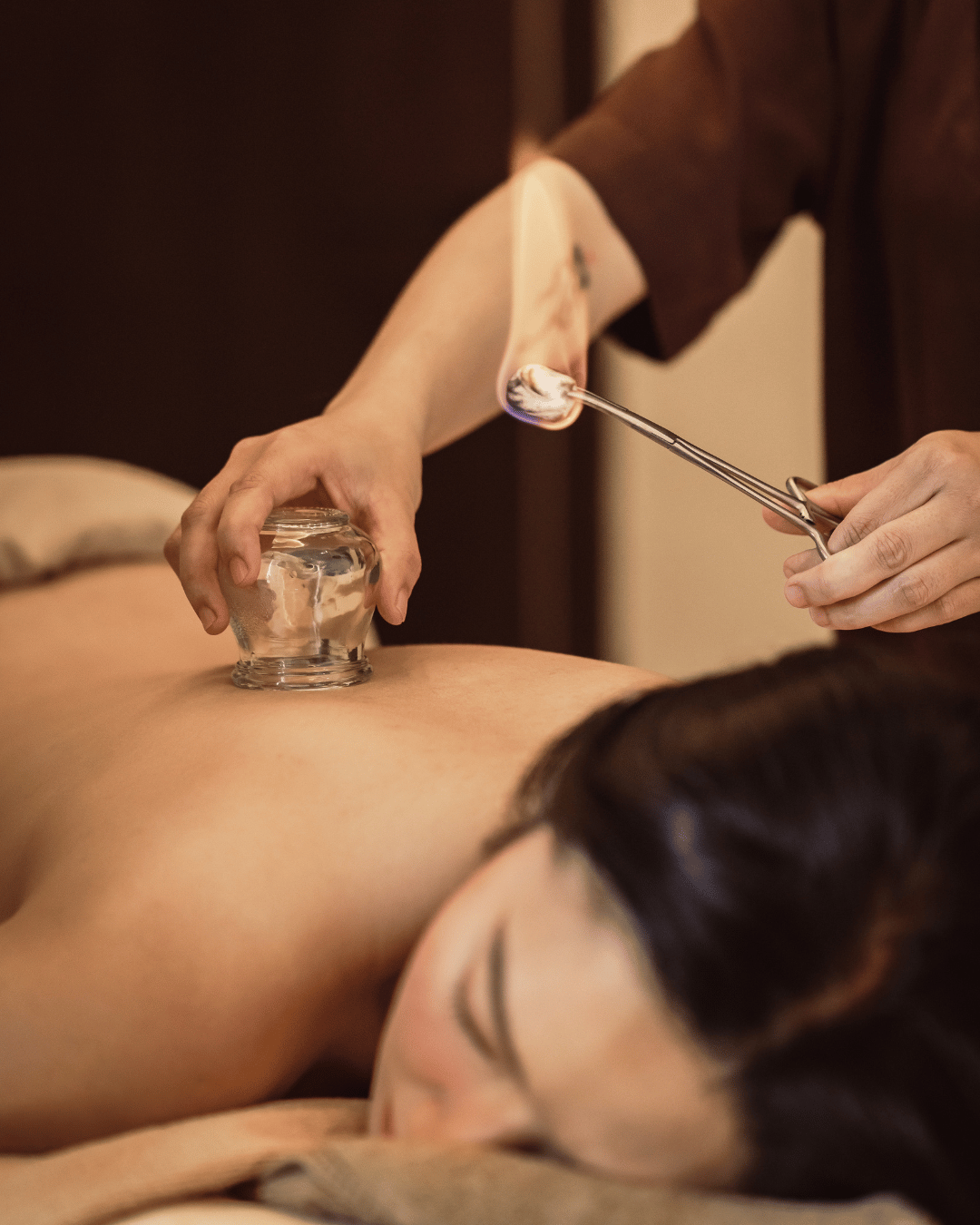 A person receiving cupping therapy on their back with multiple glass cups, while another person uses a metal tool to apply it. | Awakened Soul - Jessika Harrison 