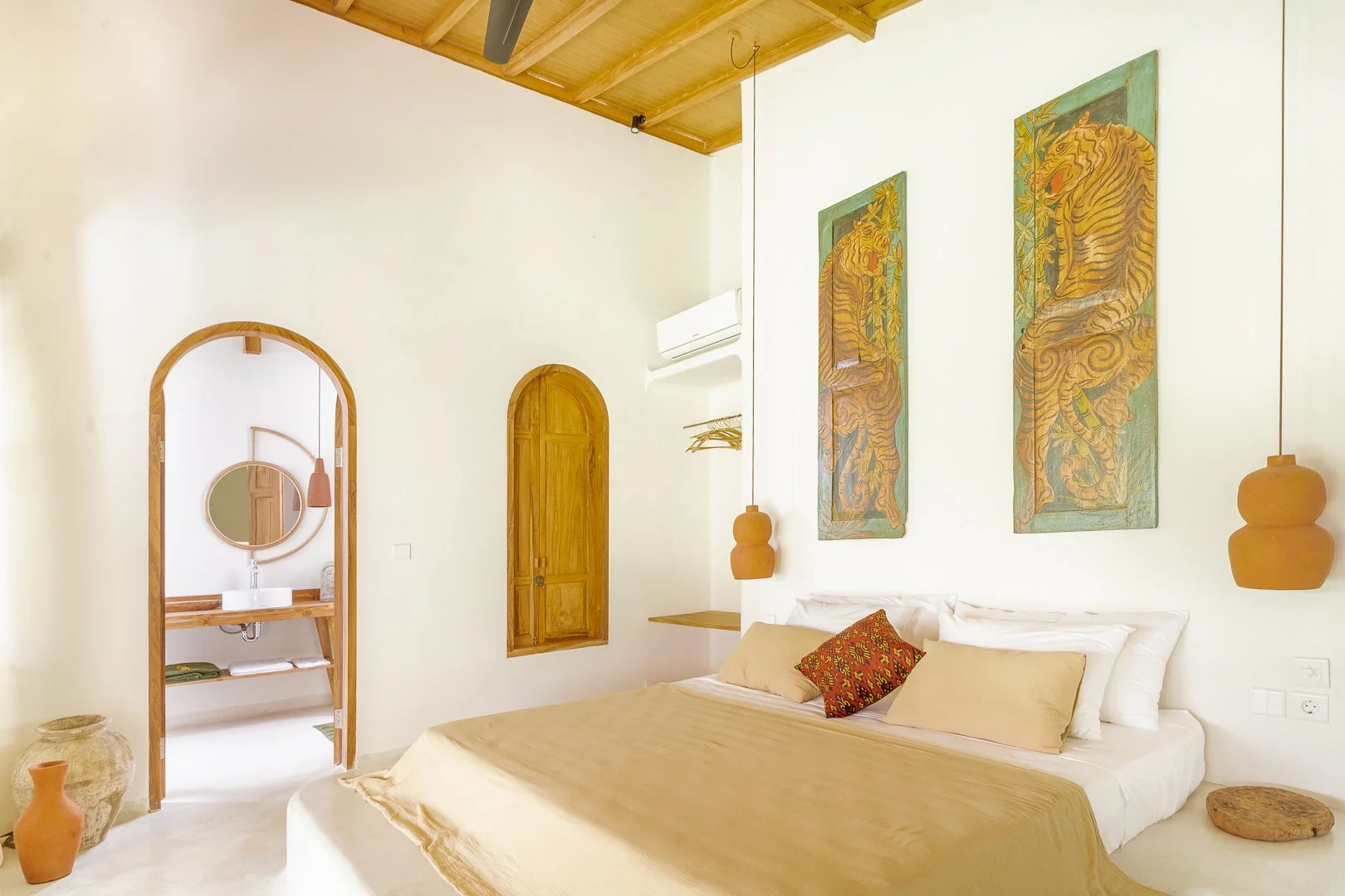 A bedroom with white walls and wooden accents, including a bed with beige and patterned pillows, artwork of tigers on the wall, and a small bathroom with a mirror and sink visible through an arched doorway.