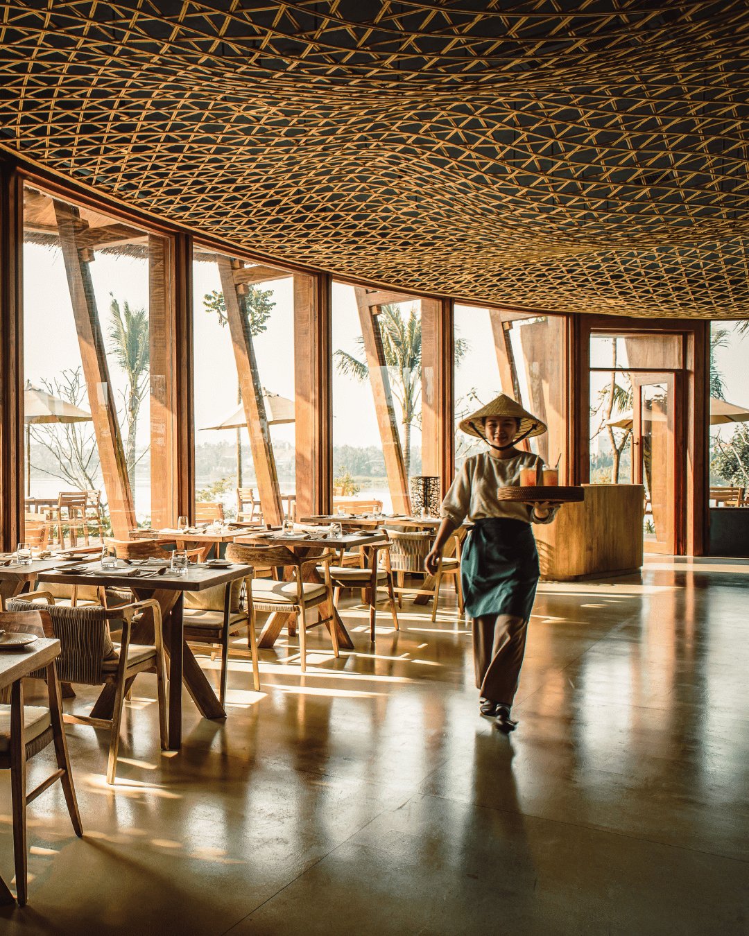 Interior of a restaurant with wooden tables and chairs, large windows with a view of palm trees outside, a woman in traditional Asian attire carrying a tray, and a decorative patterned ceiling. | Awakened Soul - Jessika Harrison 