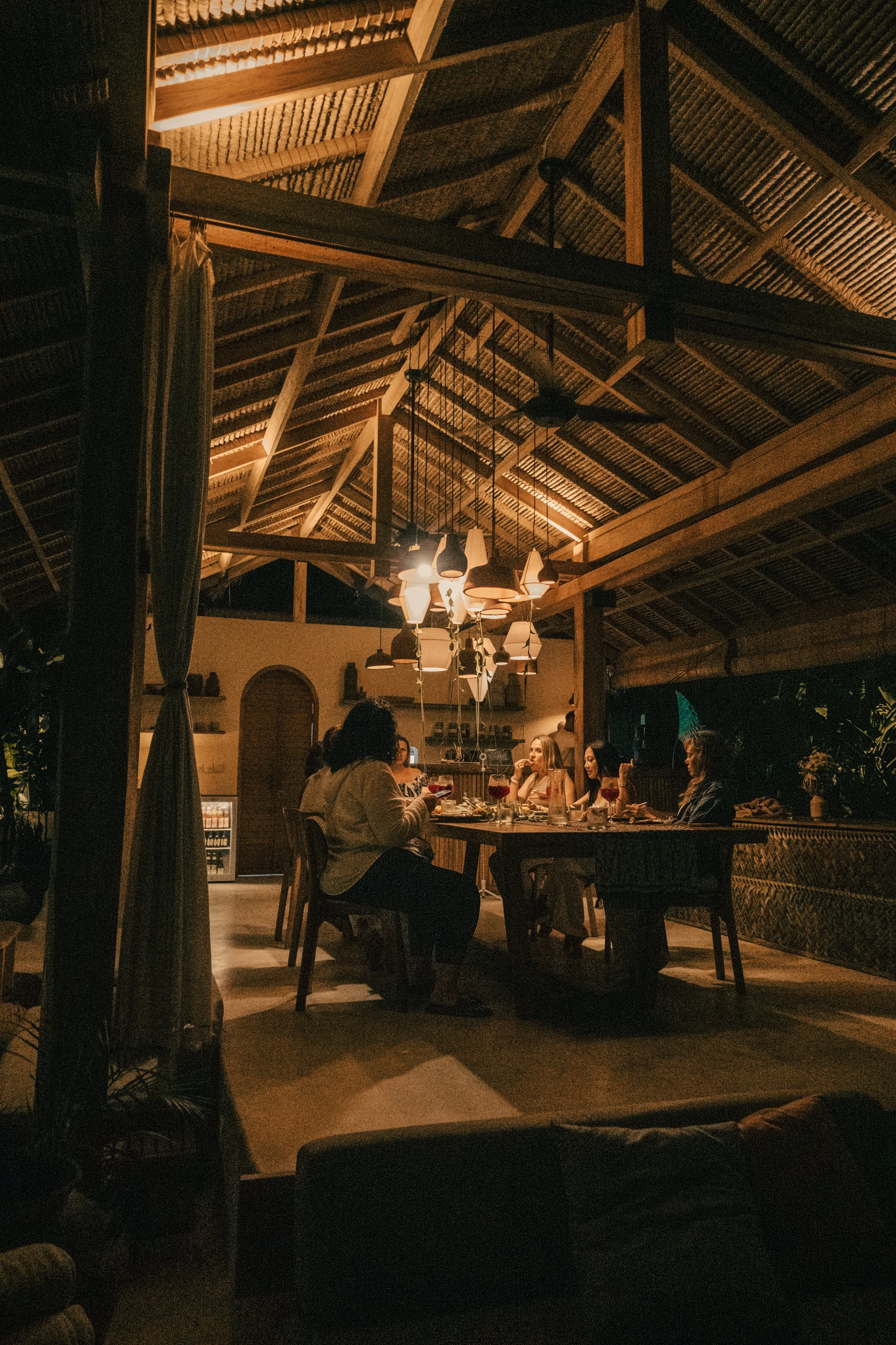 A group of people sitting at a large wooden table in a dimly lit restaurant with a high thatched ceiling and hanging pendant lights.