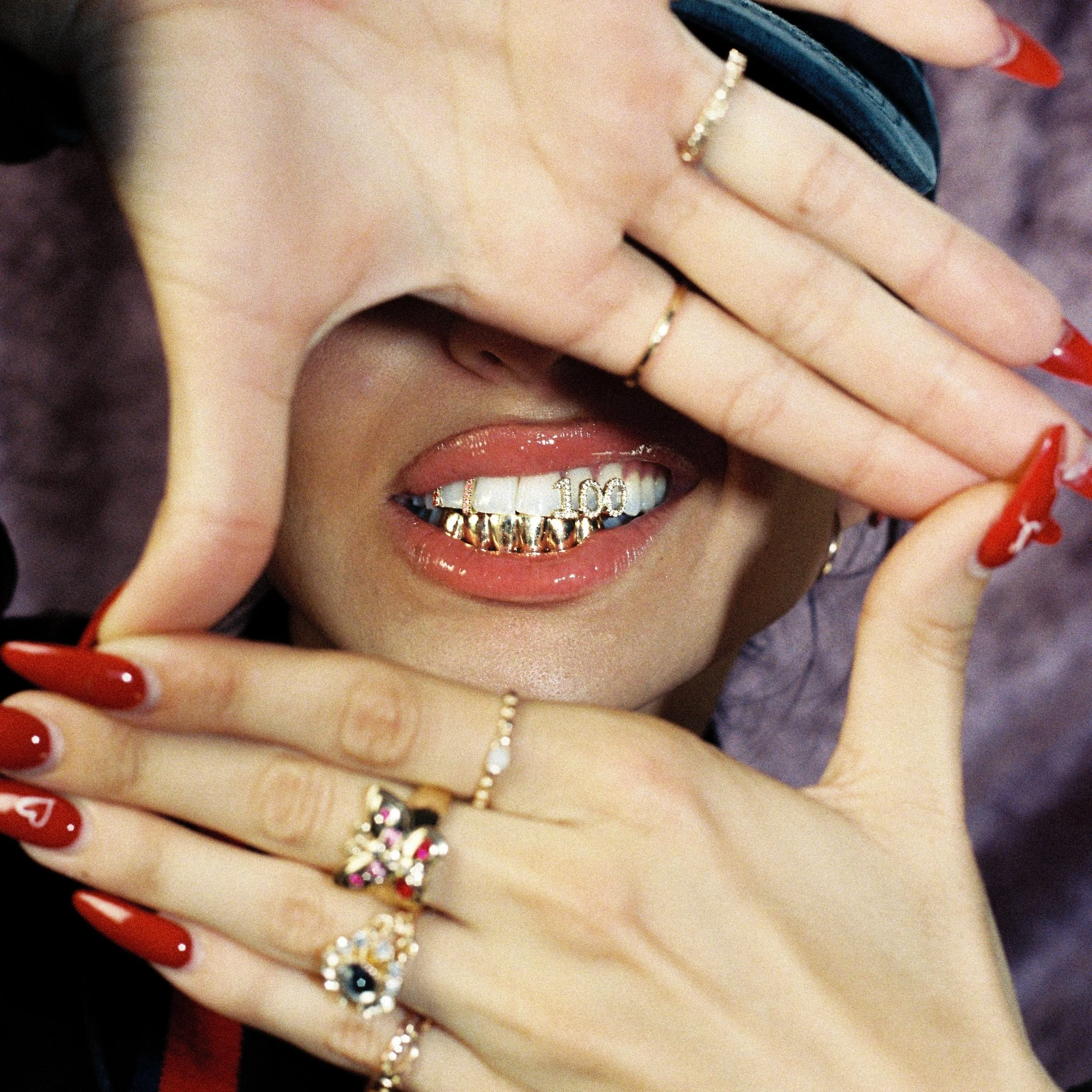 Person showing gold and diamond grillz teeth with rings and red nails, framing face with hands.