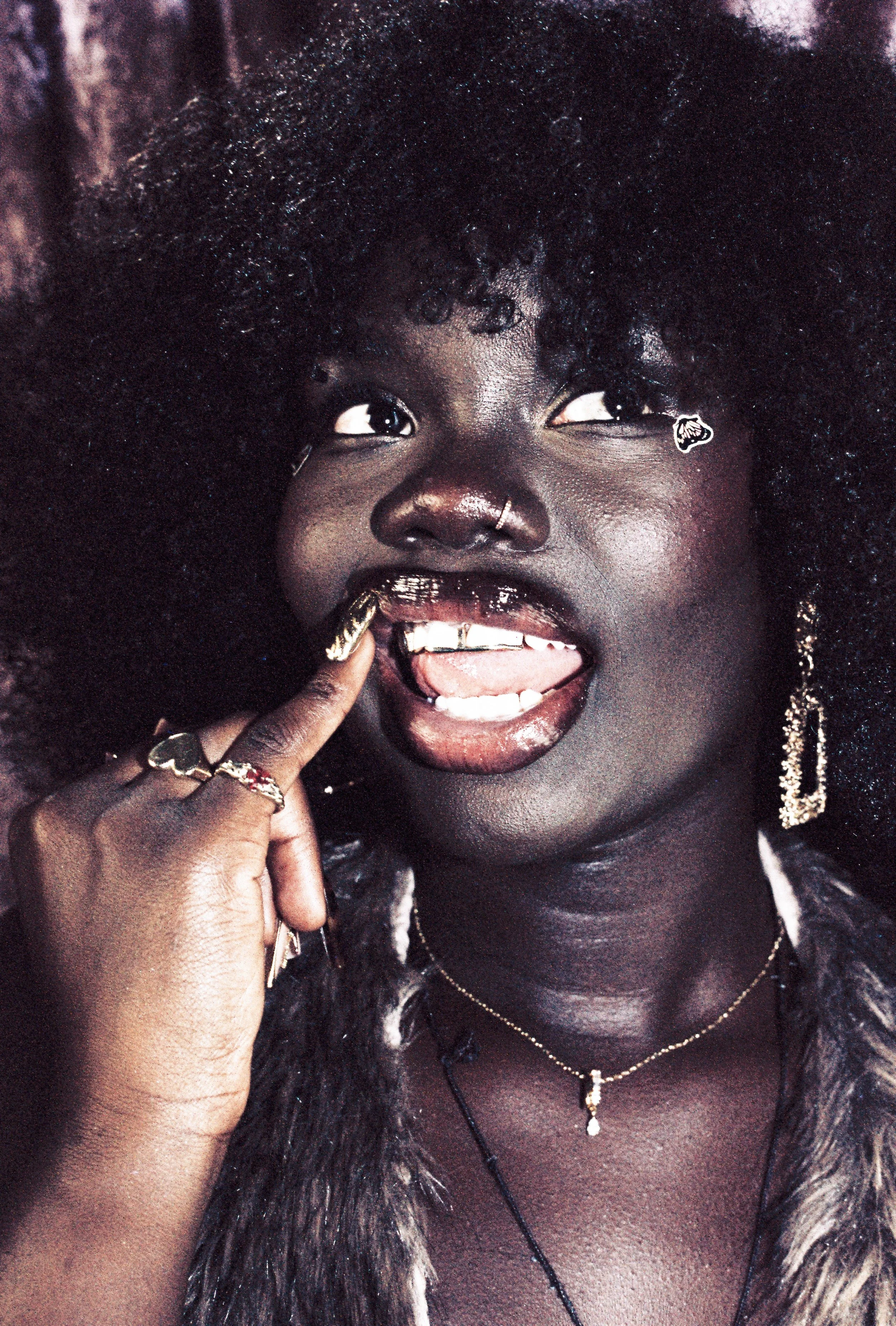Woman with curly hair, wearing jewelry and pointing at her teeth, has gold embellishments on lips, nail art, and face stickers.