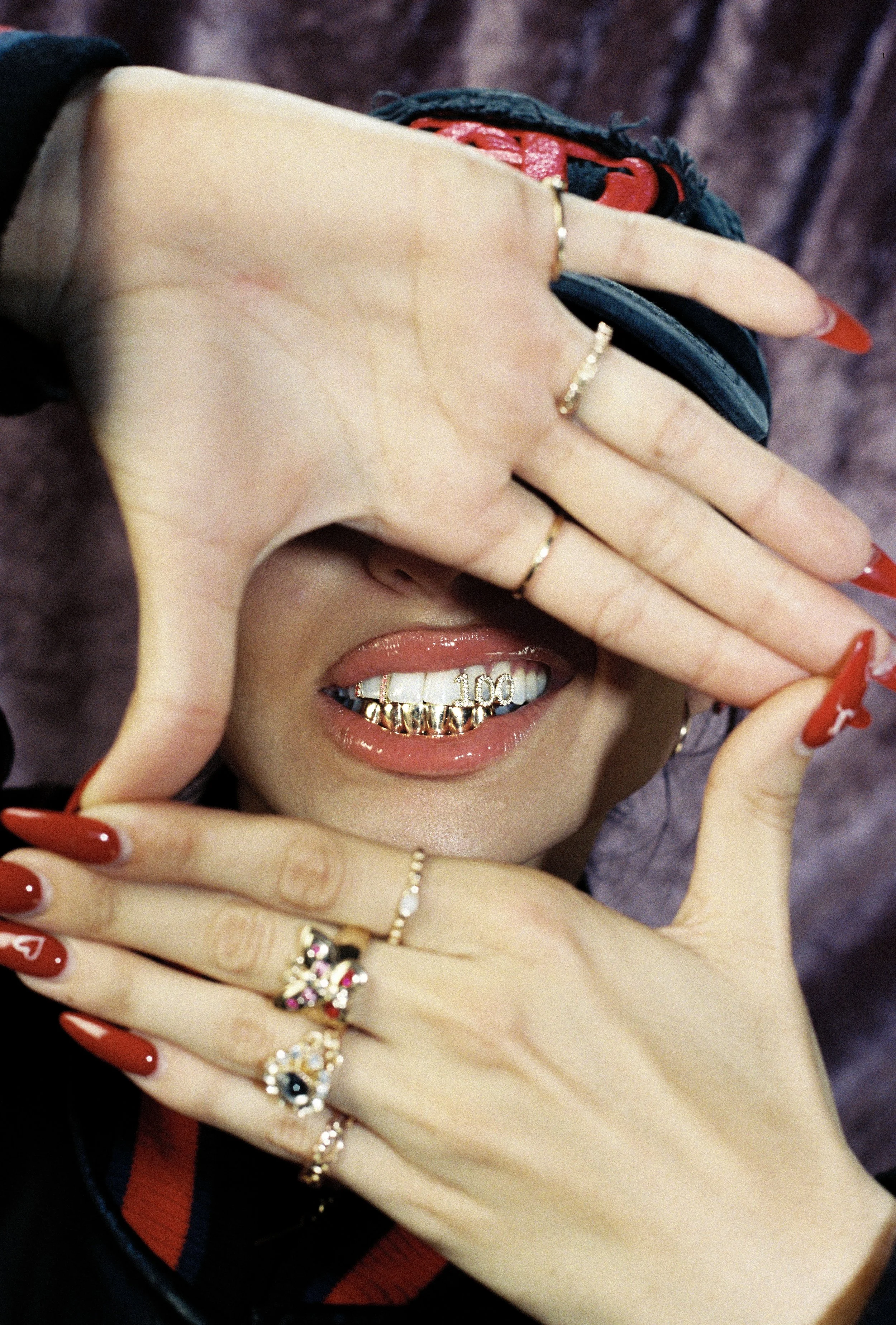 Person with long red nails, wearing multiple rings, covers part of their face with hands, revealing a smile with decorative gold grillz featuring the number "100."