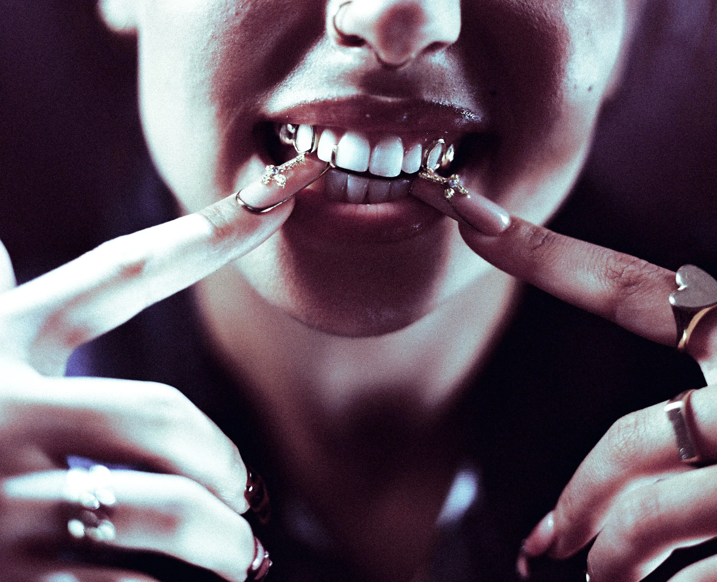 Close-up of a person smiling with gold teeth and long manicured nails, adorned with multiple rings.