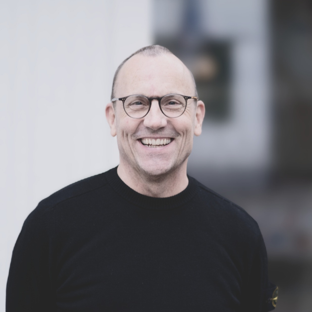 Ein lächelnder Mann mit Brille, schwarzem Pullover, kurze Haare und Glatze, vor einem unscharfen Hintergrund.
