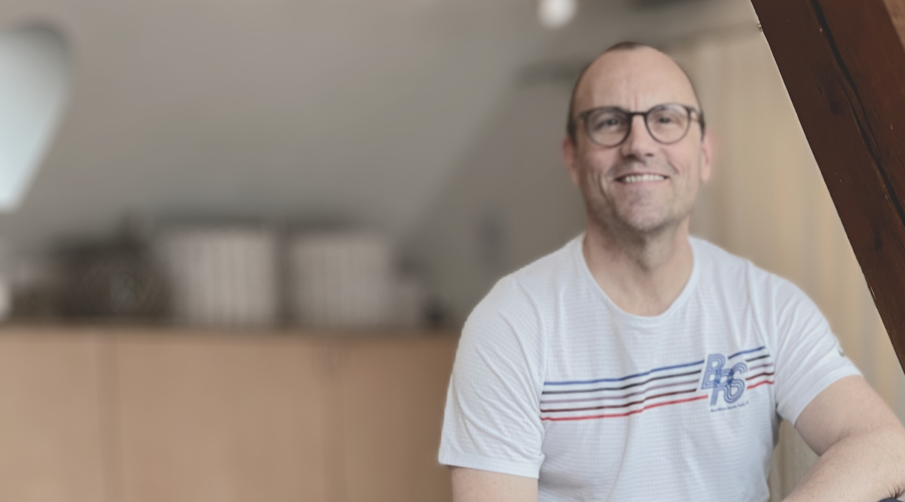 Ein lächelnder Mann mit Brille in einem weißen T-Shirt sitzt in einem Raum.