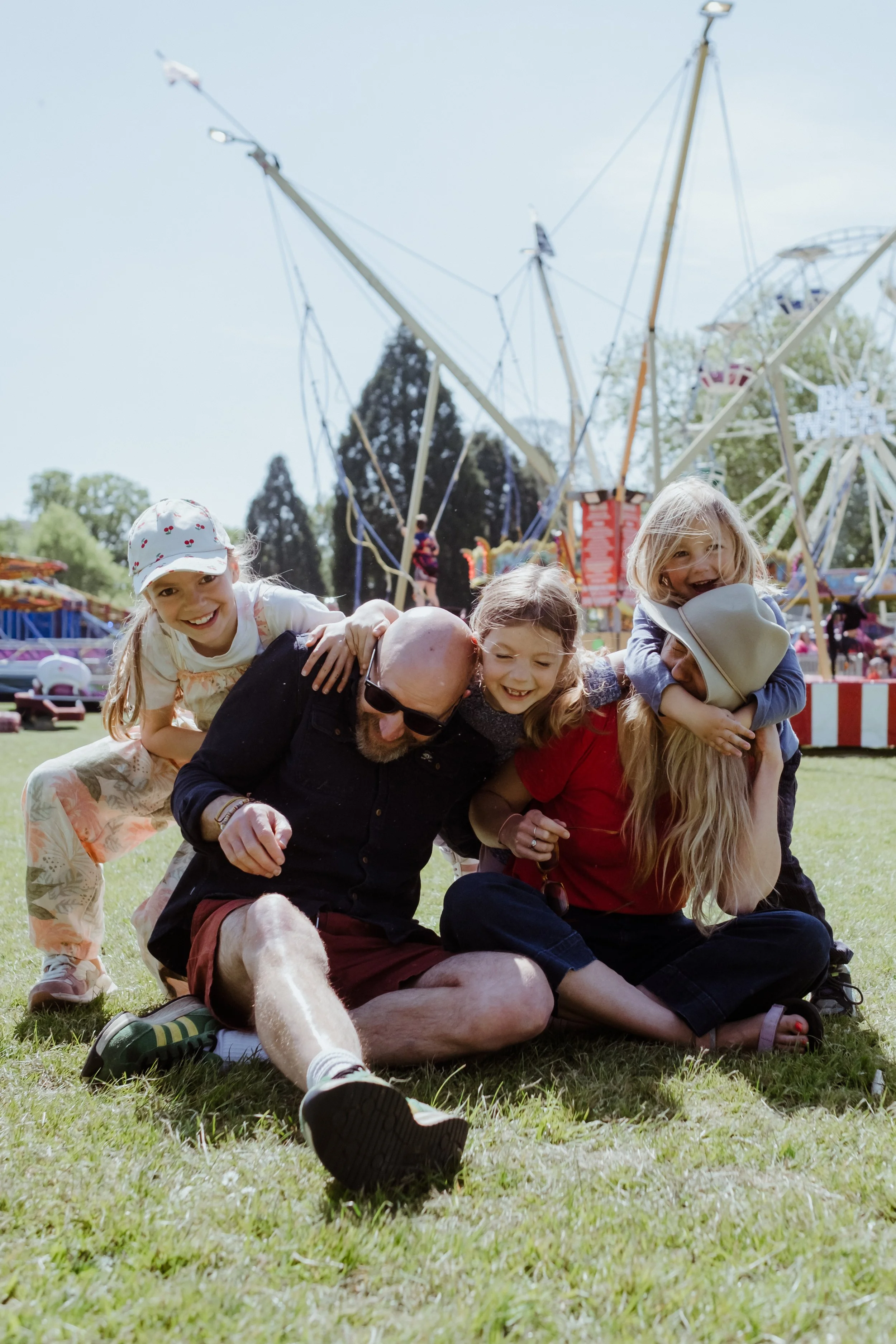 Group of children and an adult playing and smiling on the grass at a fairground with amusement rides in the background.