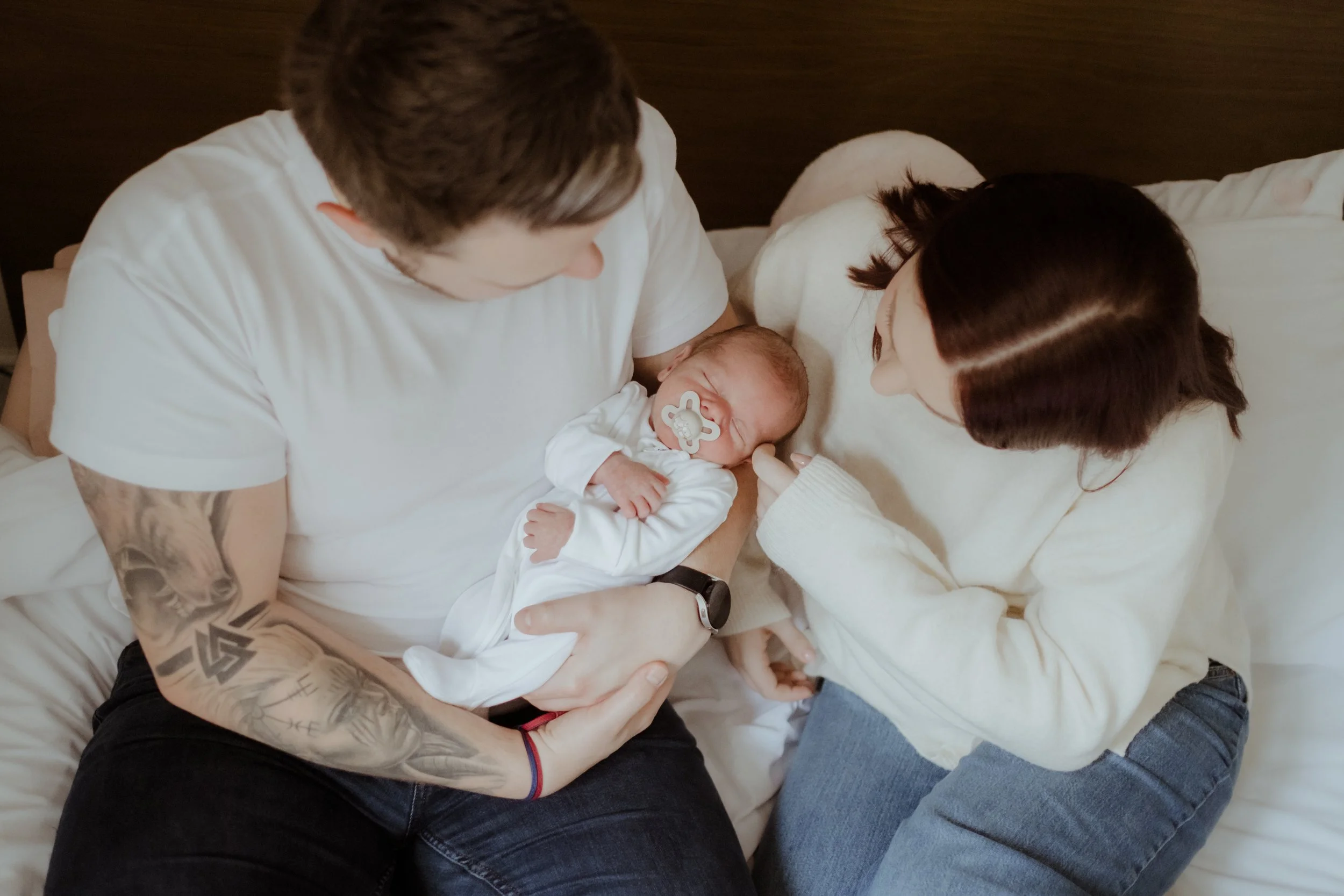 A family in a hospital room with a newborn baby. The father, with tattoos on his arm, is holding the baby and looking at the mother, who is lying in bed and gently touching the baby's hand. The scene depicts a tender moment after childbirth.