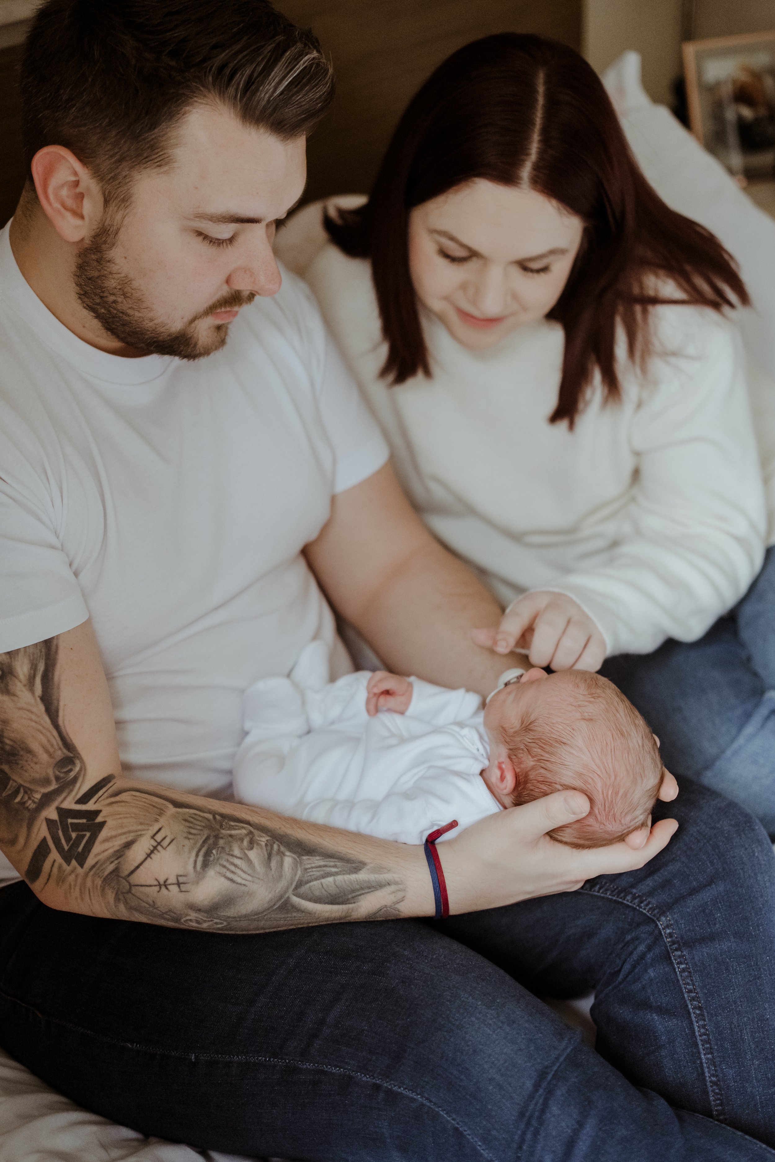 A man with tattoos is holding a newborn baby, with both adults sitting close together on a bed. A woman next to them is looking at the baby with a smile.