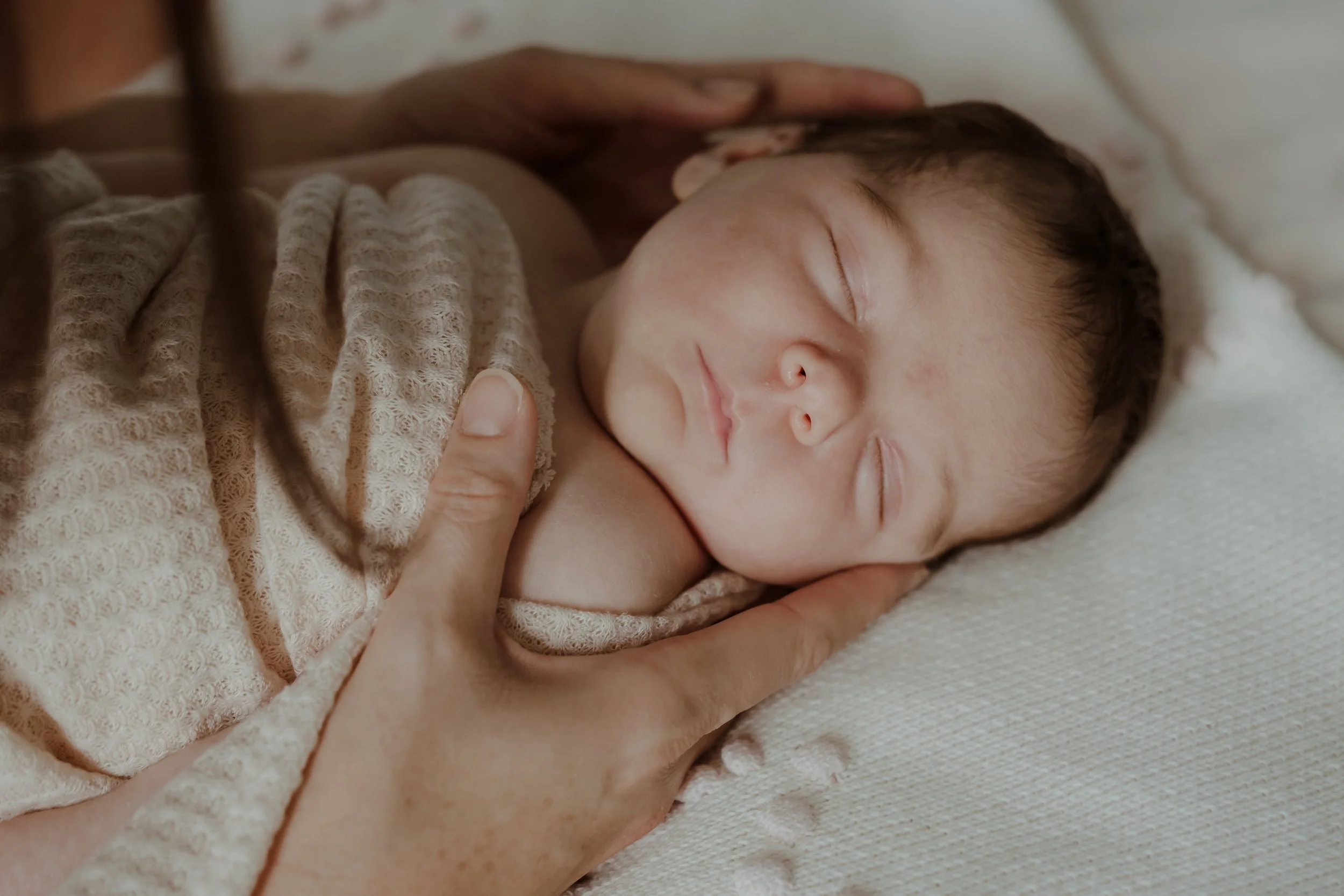 Close-up of a newborn baby sleeping, being gently held by an adult, wrapped in a beige knitted blanket.