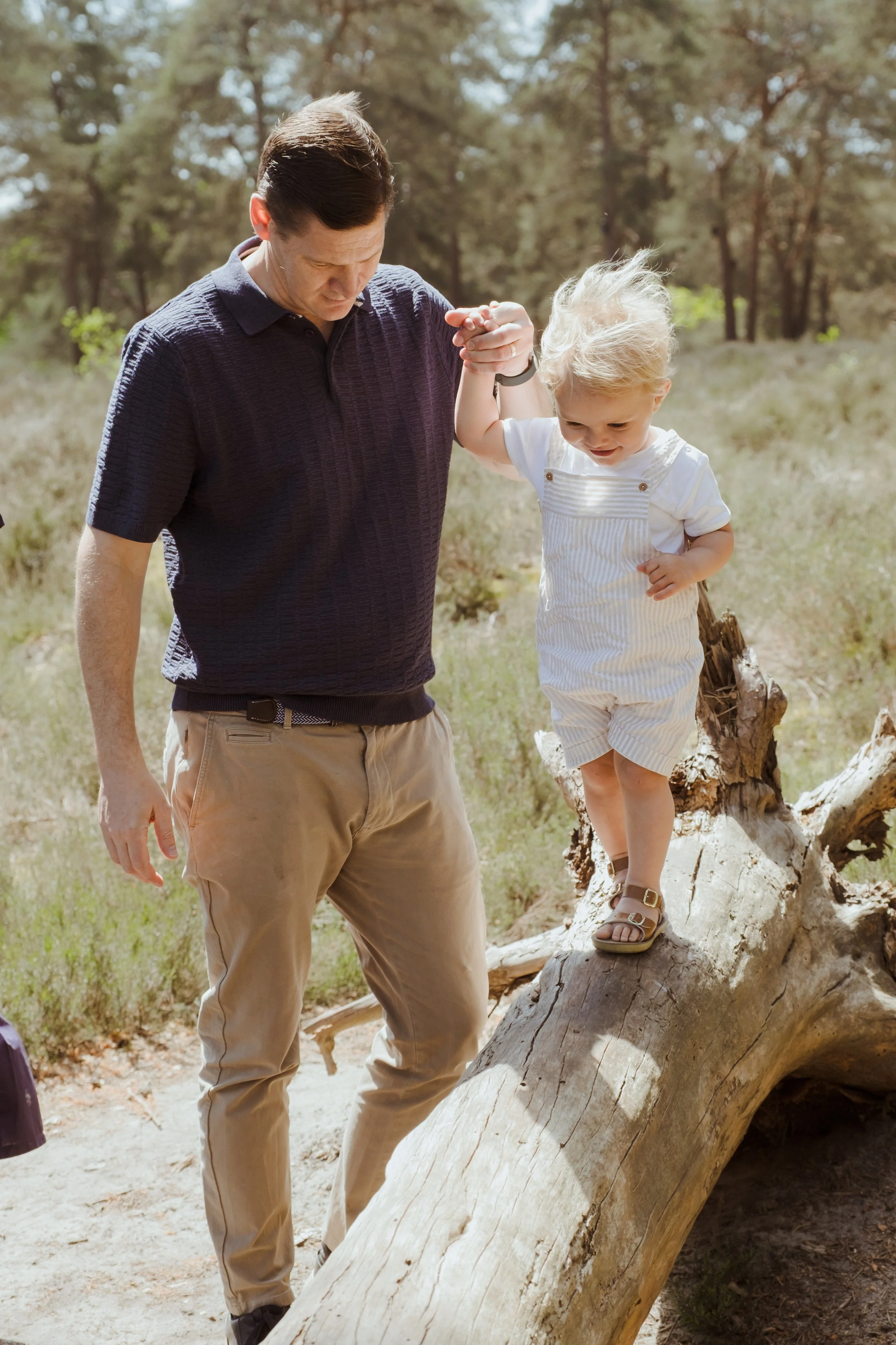 A man helping a young girl walk across a fallen tree in a forested area during daytime.