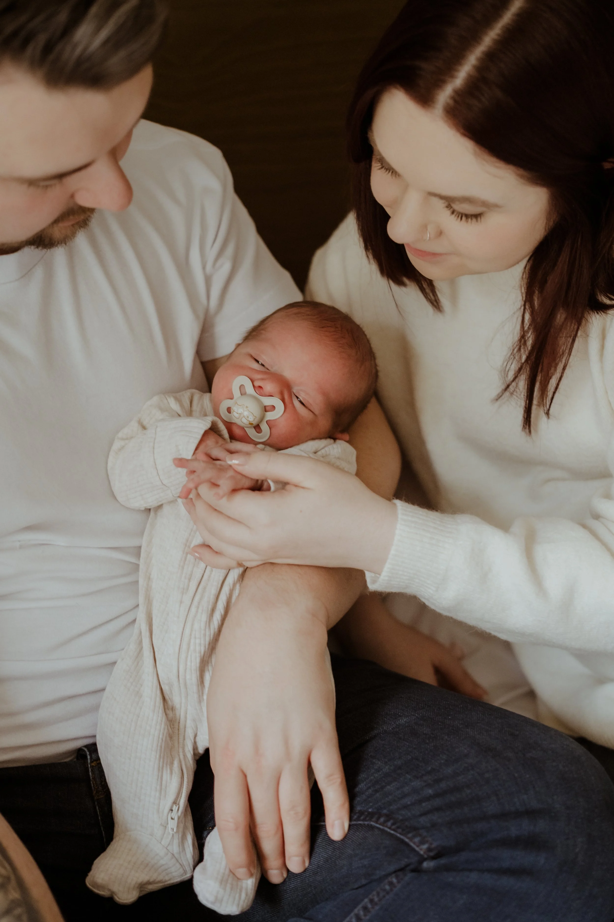 A man and a woman holding a newborn baby together, with the man on the left and the woman on the right, both looking at the baby with affection. The baby has a pacifier in its mouth.