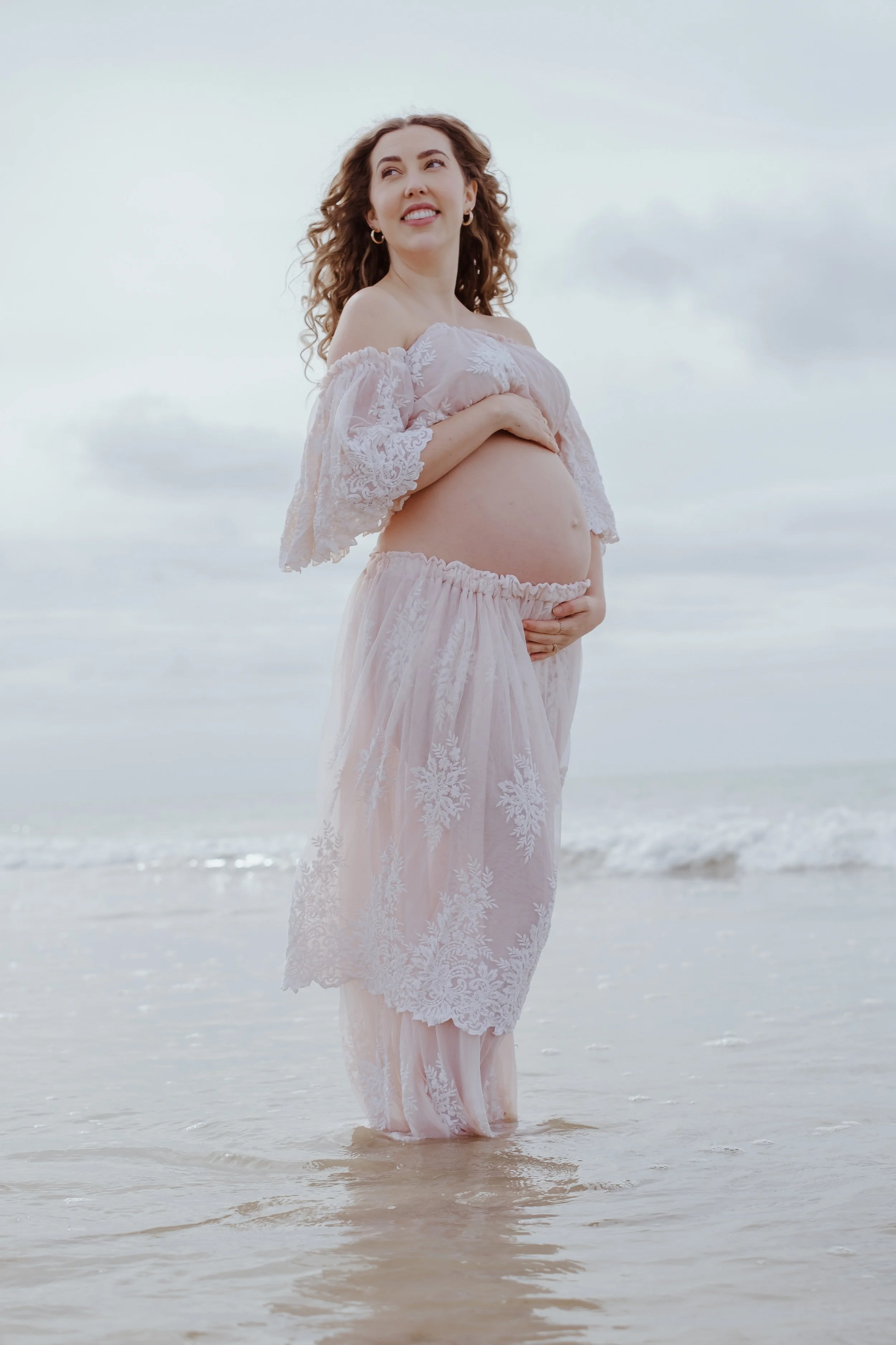 A pregnant woman wearing a light pink, lace, off-shoulder dress stands in shallow water at the beach, smiling and looking off to the side. Her left hand rests on her belly, and her right hand supports her belly from below.