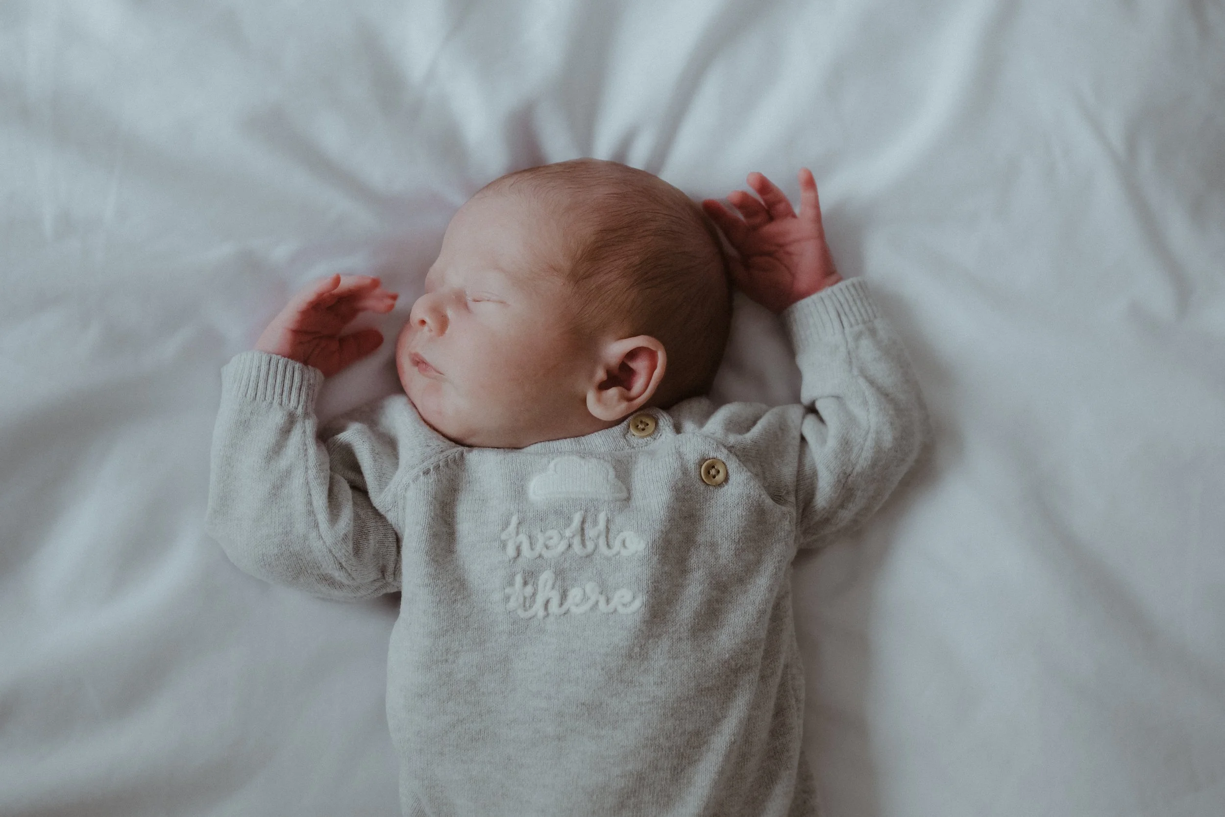 A sleeping baby with a light complexion, lying on a white bed, dressed in a gray onesie with buttons and the words 'hello there' embroidered on it.