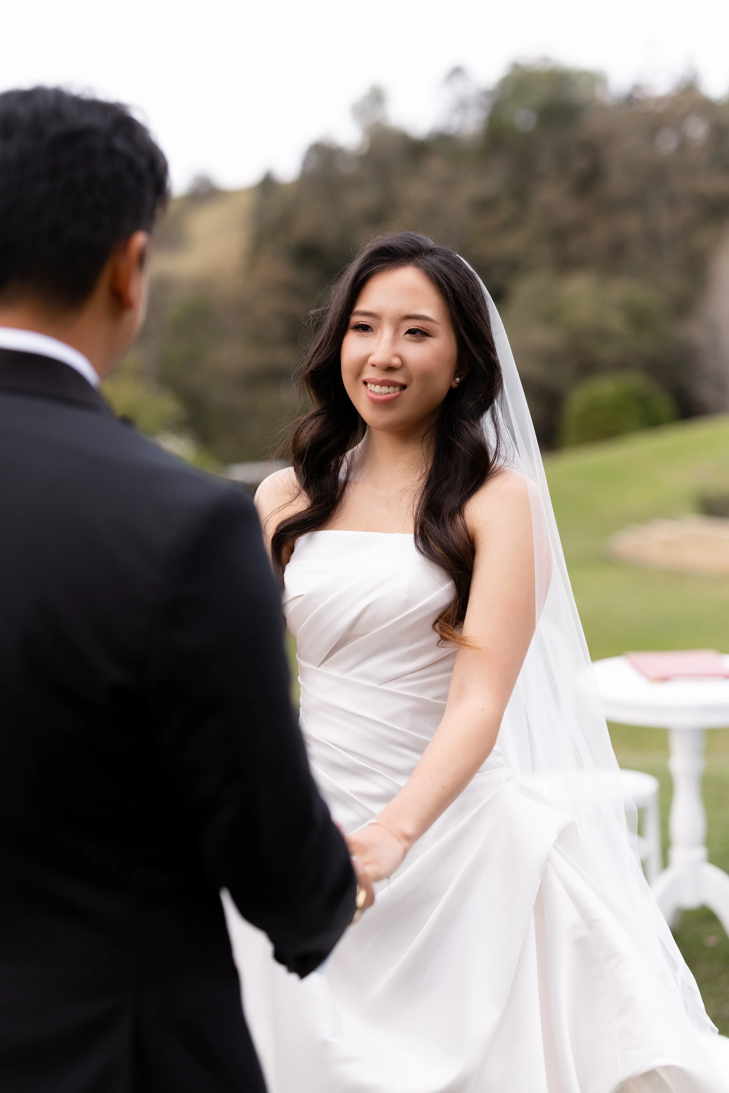 Alicia smiling in her gown and veil, holding hands with Ezra on the lawn