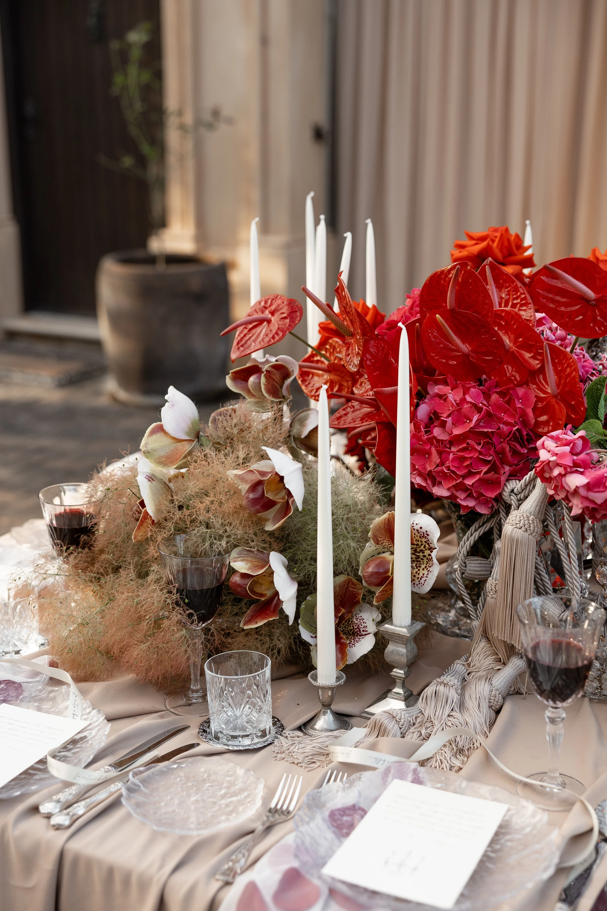 Elegant dining table decorated with floral arrangements including red and pink flowers, tall white candles, and glassware set on a cream-colored tablecloth.