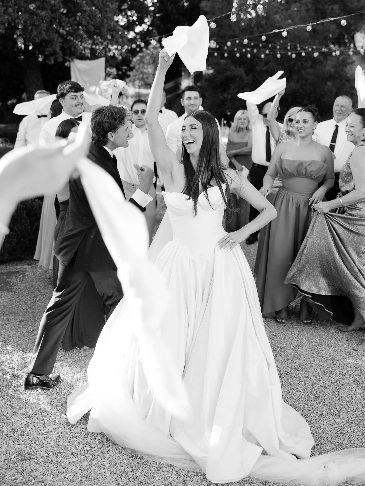 Black and white photo of Marilena and Joshua dancing with guests waving napkins under string lights