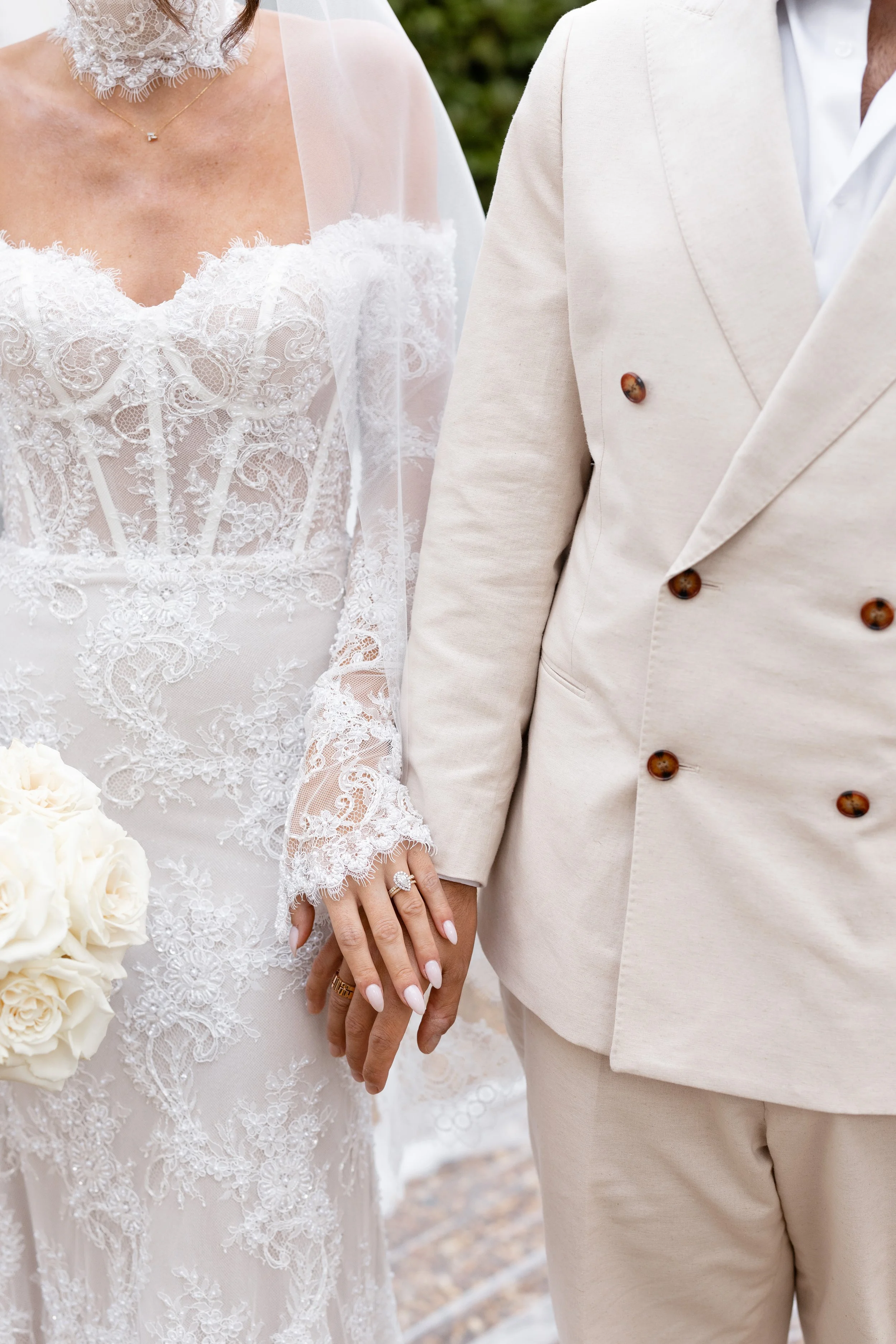 Steph and Alex holding hands, close-up of her lace bodice and his cream suit jacket