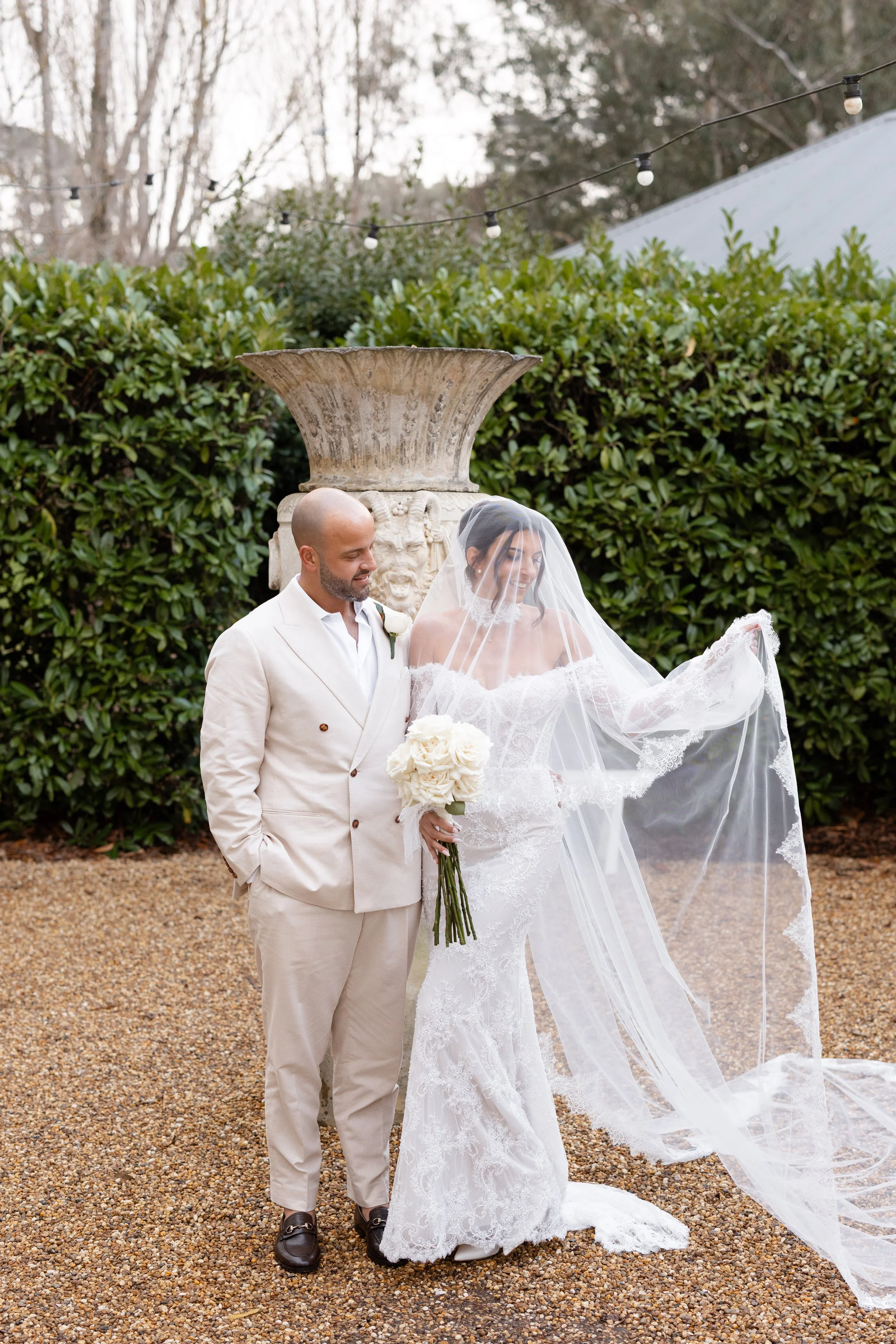 Steph and Alex under her veil beside a stone urn and hedges at Lindenderry Red Hill