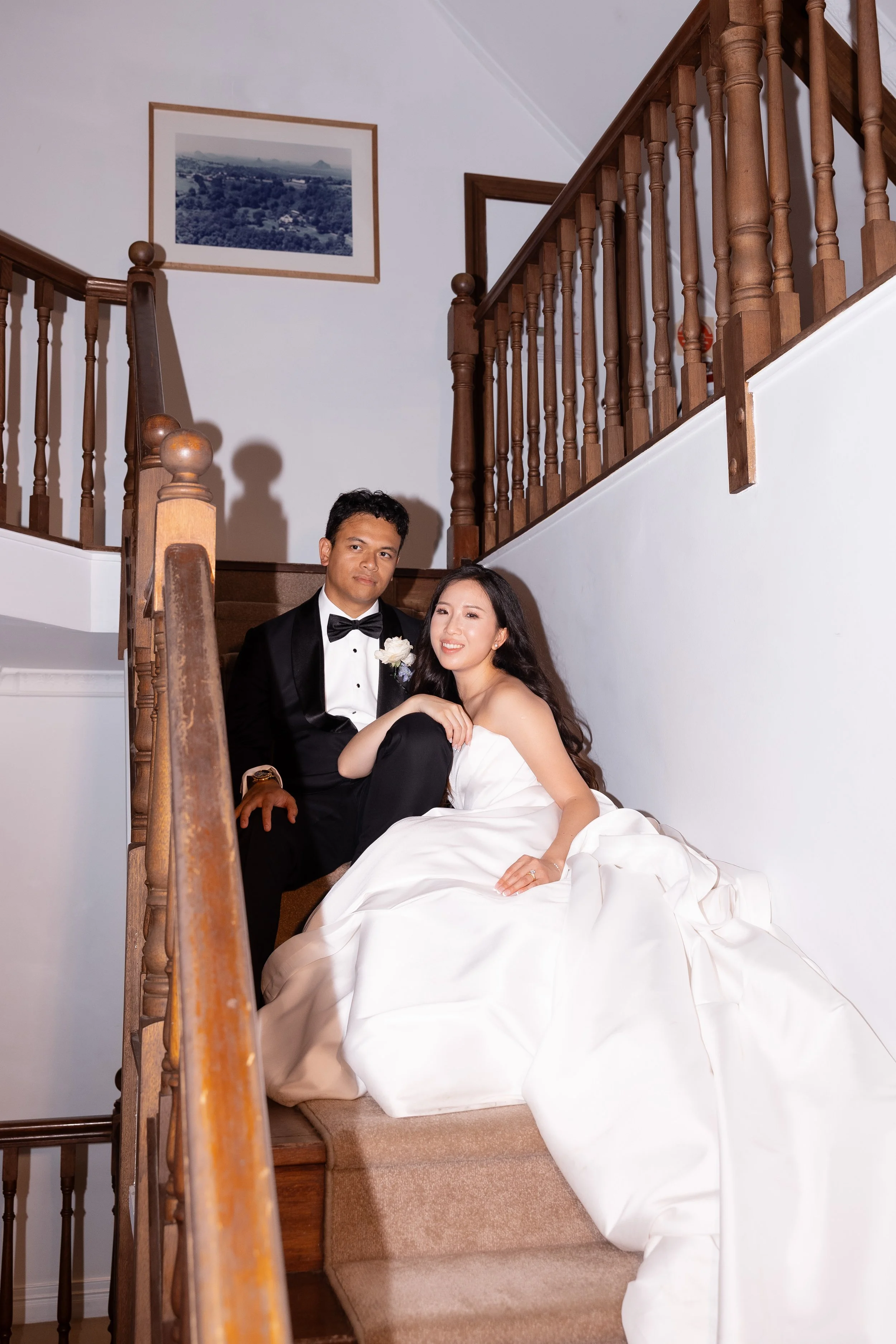 Alicia and Ezra seated together on a wooden staircase, Ezra in a black suit and Alicia in a white gown