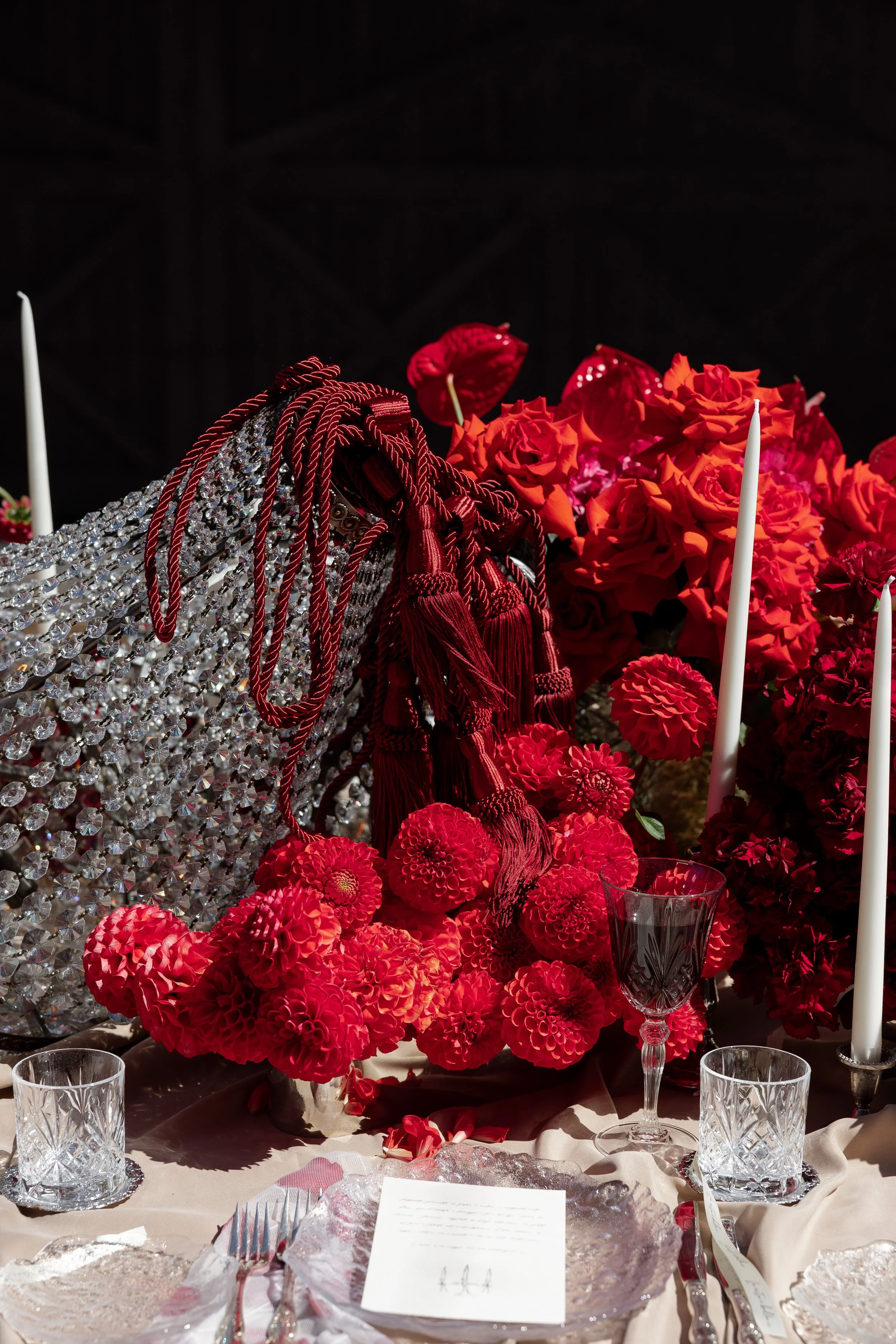 A table decorated with a large floral arrangement of red and pink flowers, red tassels, and crystal ornaments. There are white candles, crystal glasses, and cutlery on the table setting.