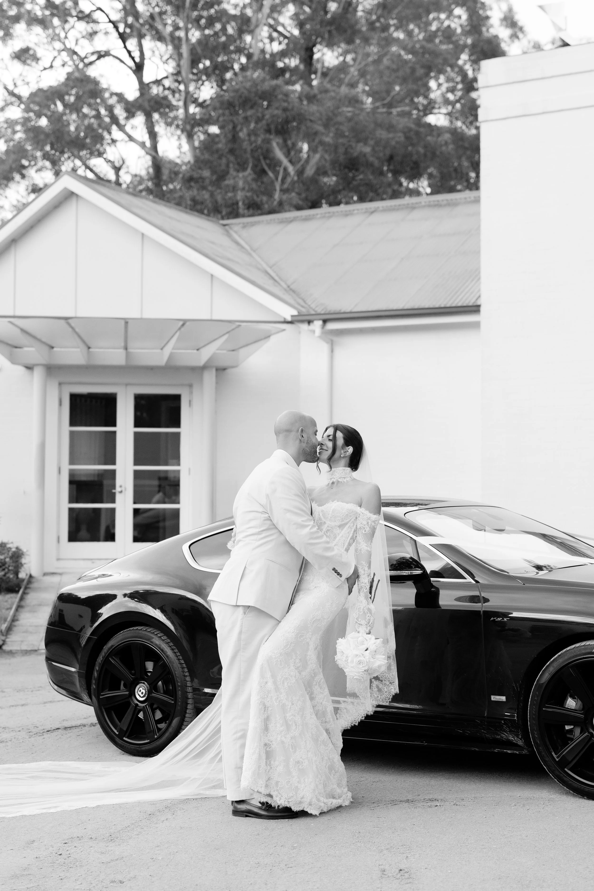 Steph and Alex beside a black Bentley by a white building at Lindenderry Red Hill, in black and white