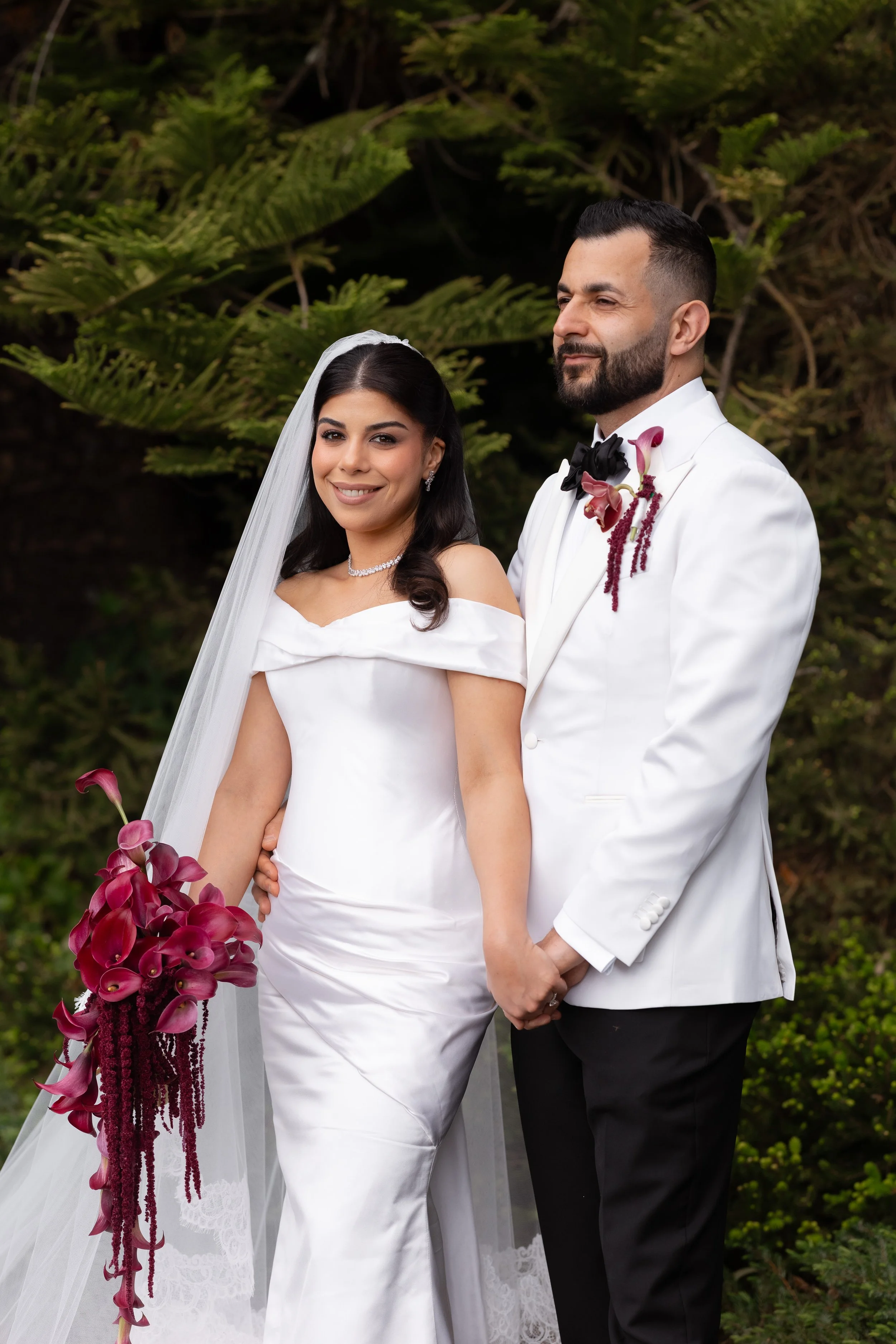 Jihan and Rob in white, posing close with her burgundy bouquet