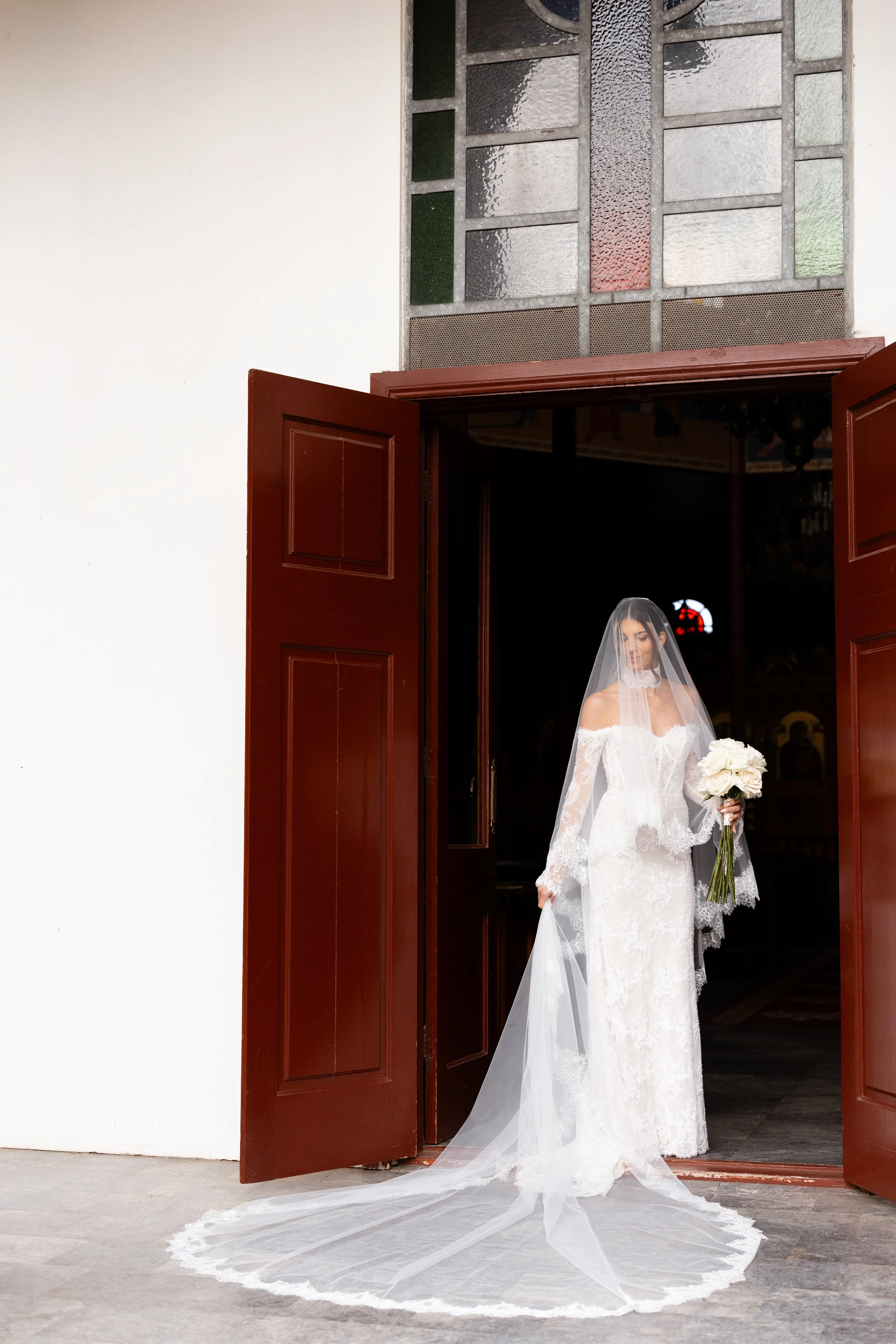 Steph in her veil and lace gown stepping out of the church doors holding a white bouquet