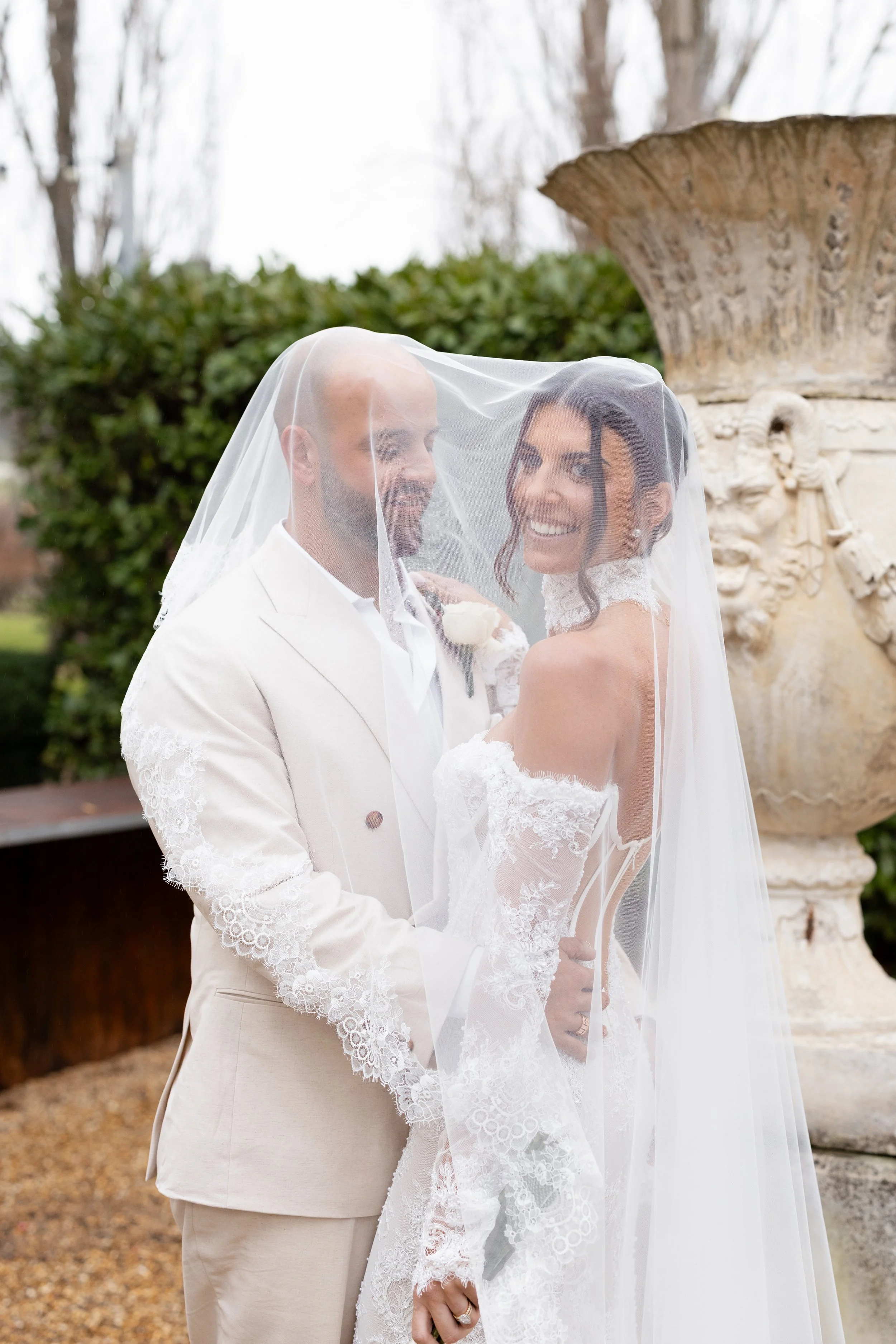 Steph and Alex smile together under her veil beside a stone urn at Lindenderry Red Hill