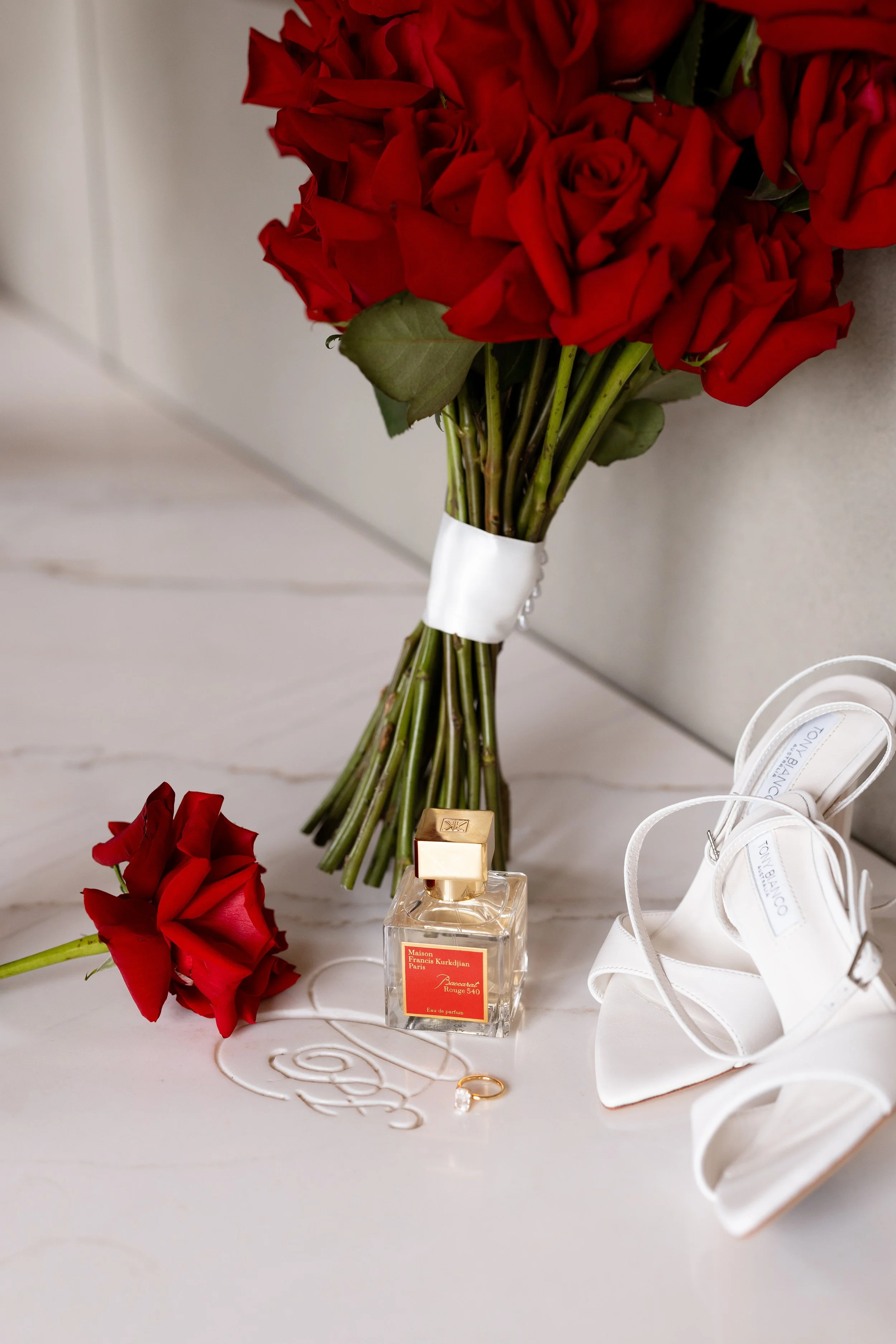 A bouquet of red roses with green leaves, a box of perfume, a white ribbon, a gold ring with a diamond, and a pair of white high-heeled sandals arranged on a white surface.