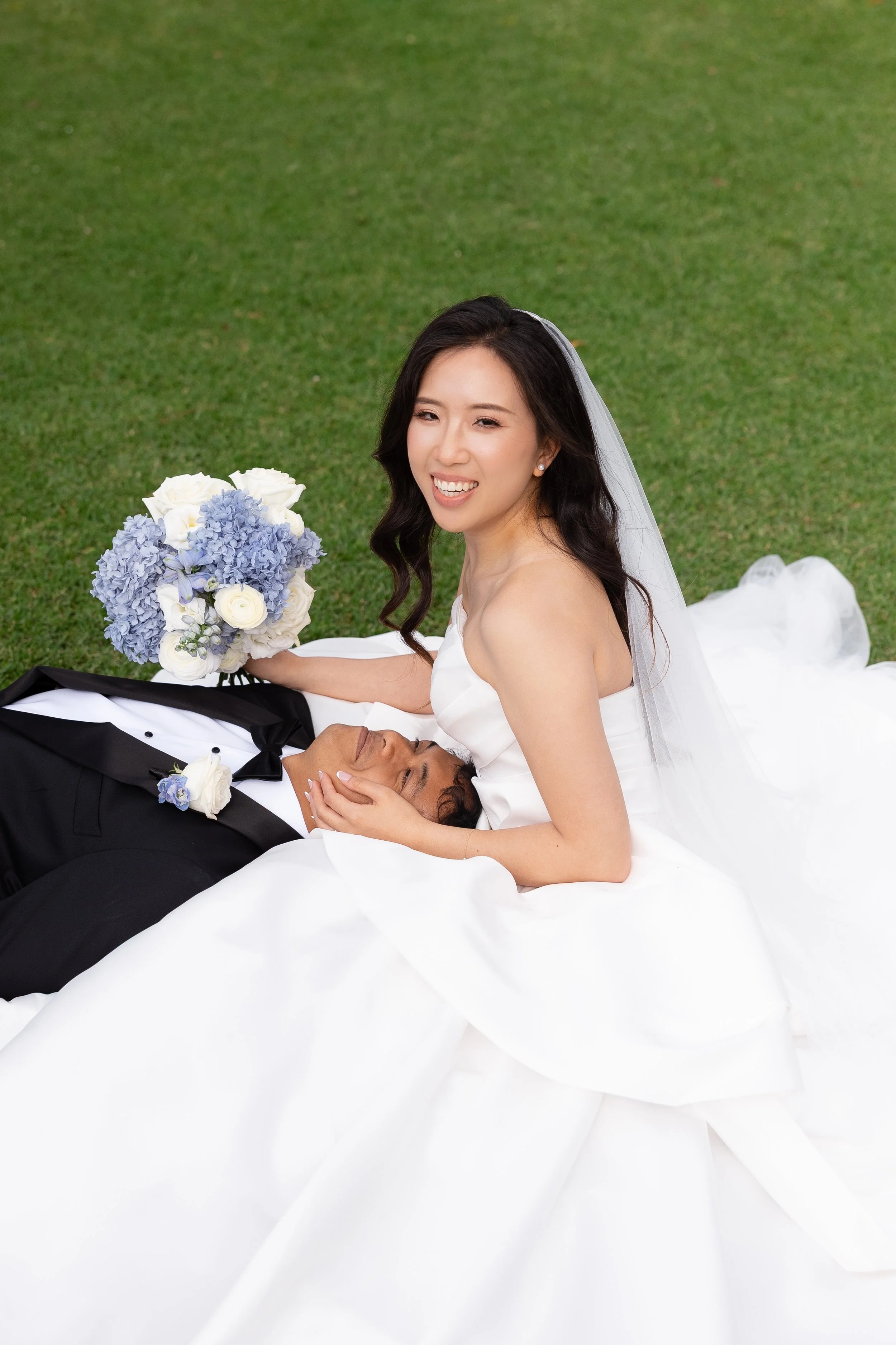Alicia sitting on the lawn smiling with a blue and white bouquet while Ezra lies on the grass