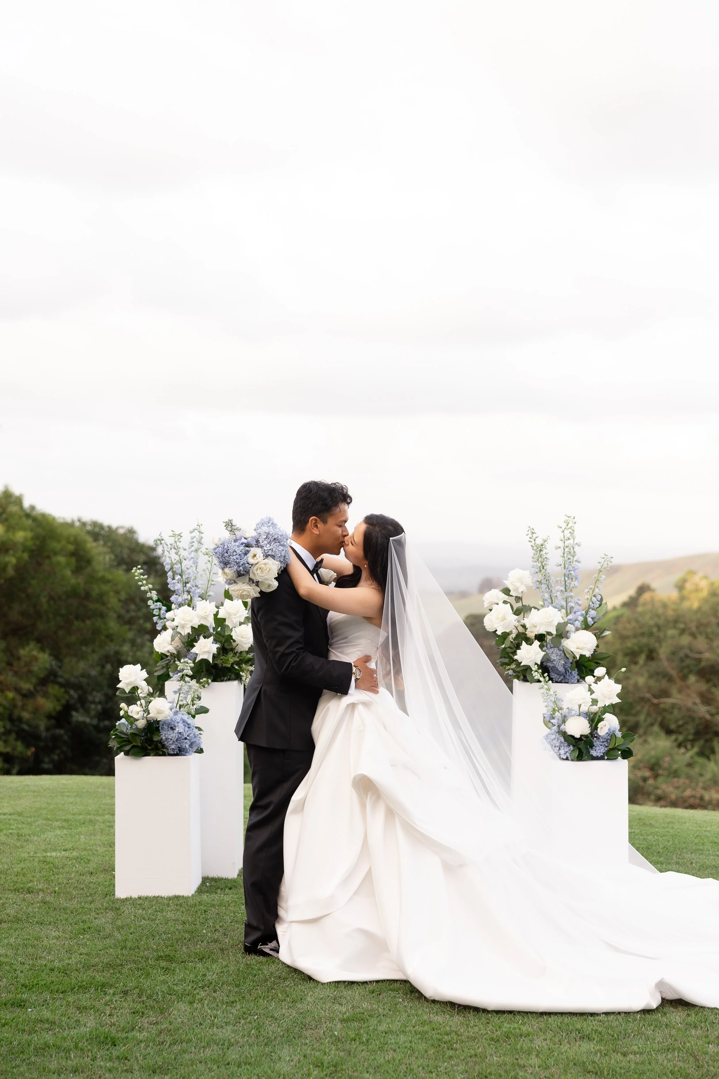 Alicia and Ezra kissing on the lawn between white and blue floral arrangements