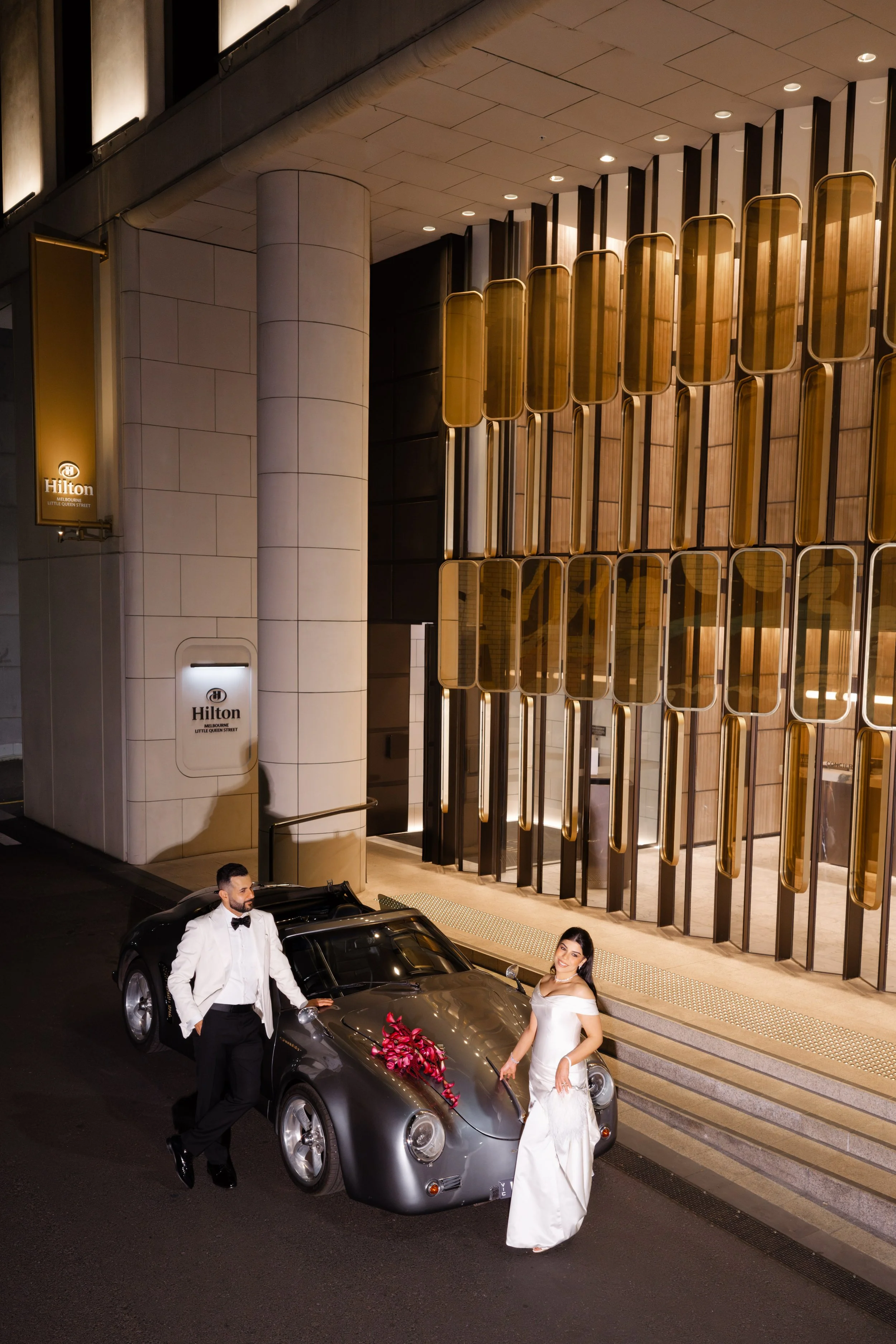 Jihan and Rob beside a classic silver convertible outside the hotel entrance