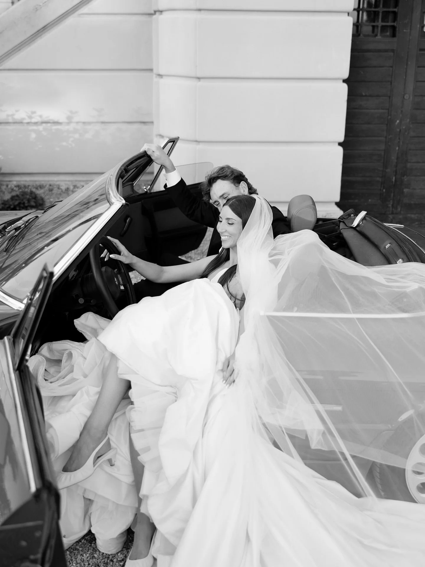 Black and white photo of Marilena and Joshua in a vintage convertible, her veil blowing over the car