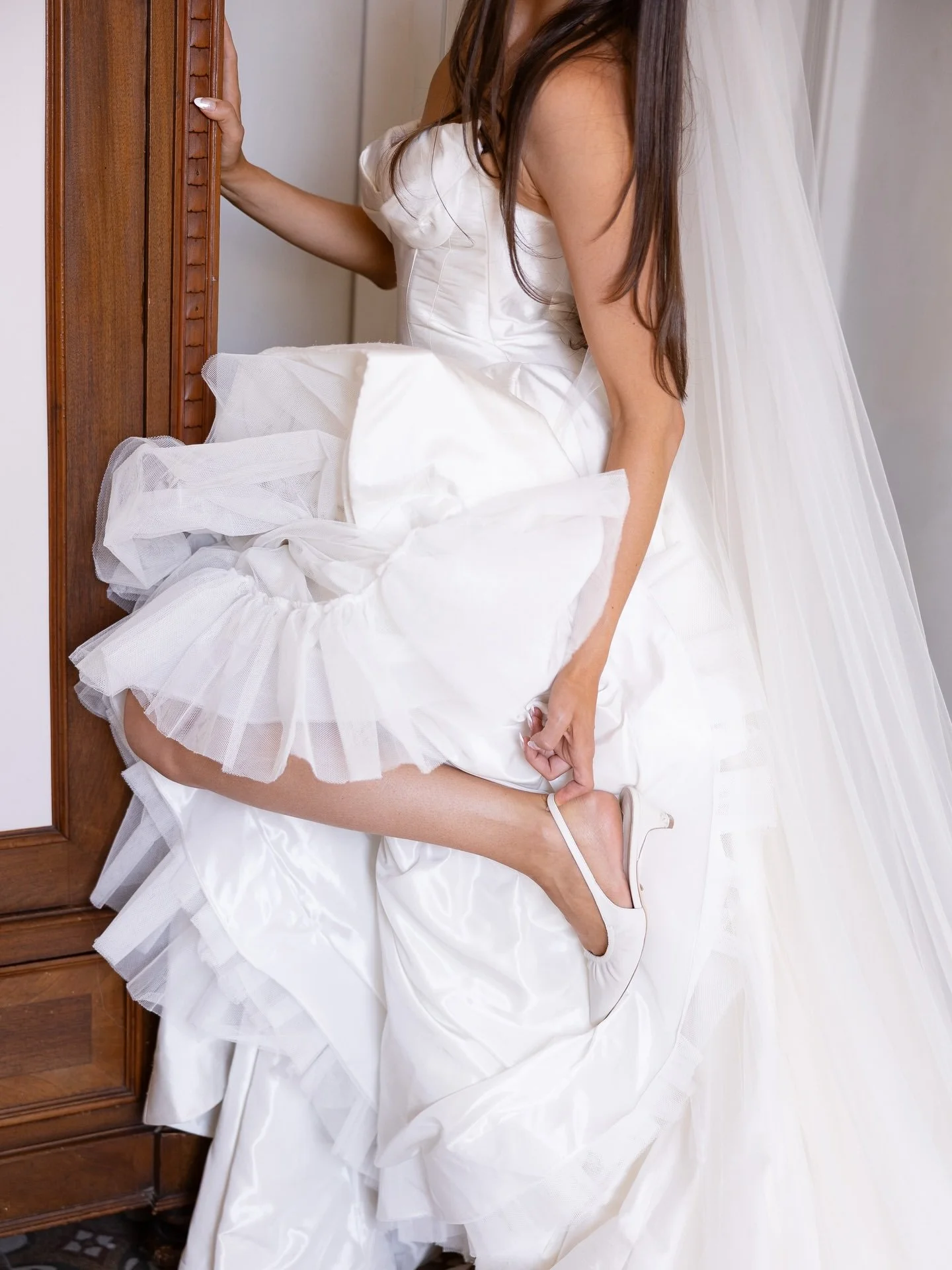 Marilena lifting the tulle skirt of her gown beside a wooden cabinet to show her wedding shoe