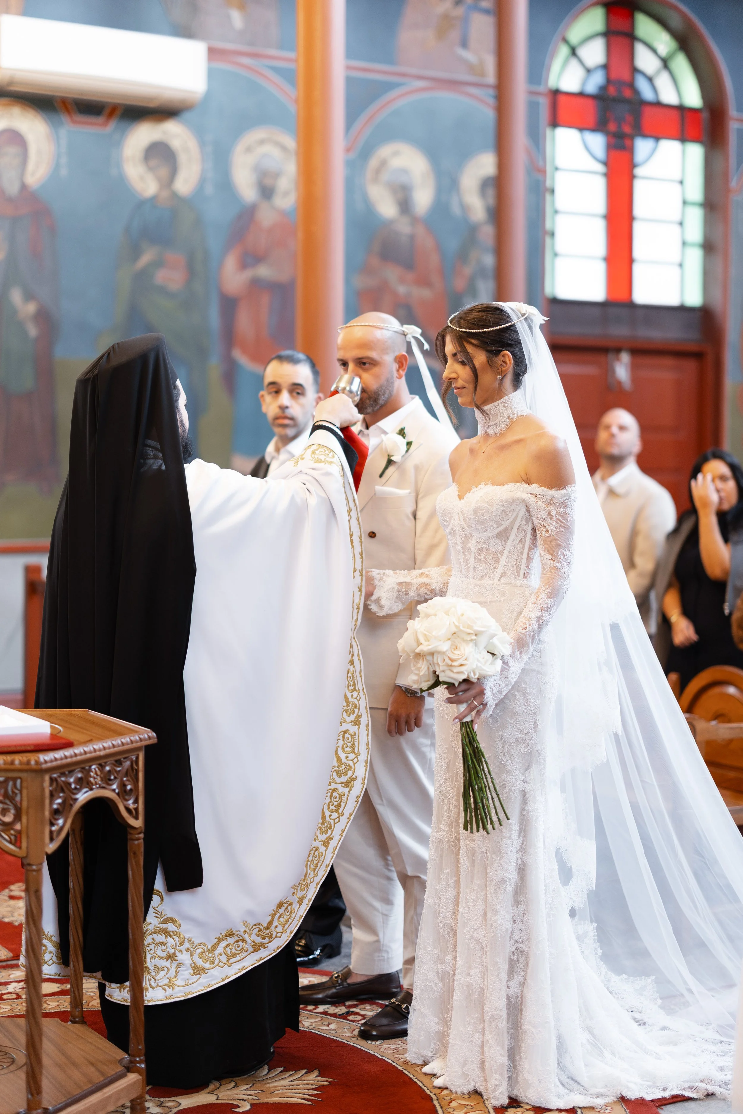 Steph and Alex with a priest holding a white cloth during the ceremony at Holy Monastery of Panagia Kamariani