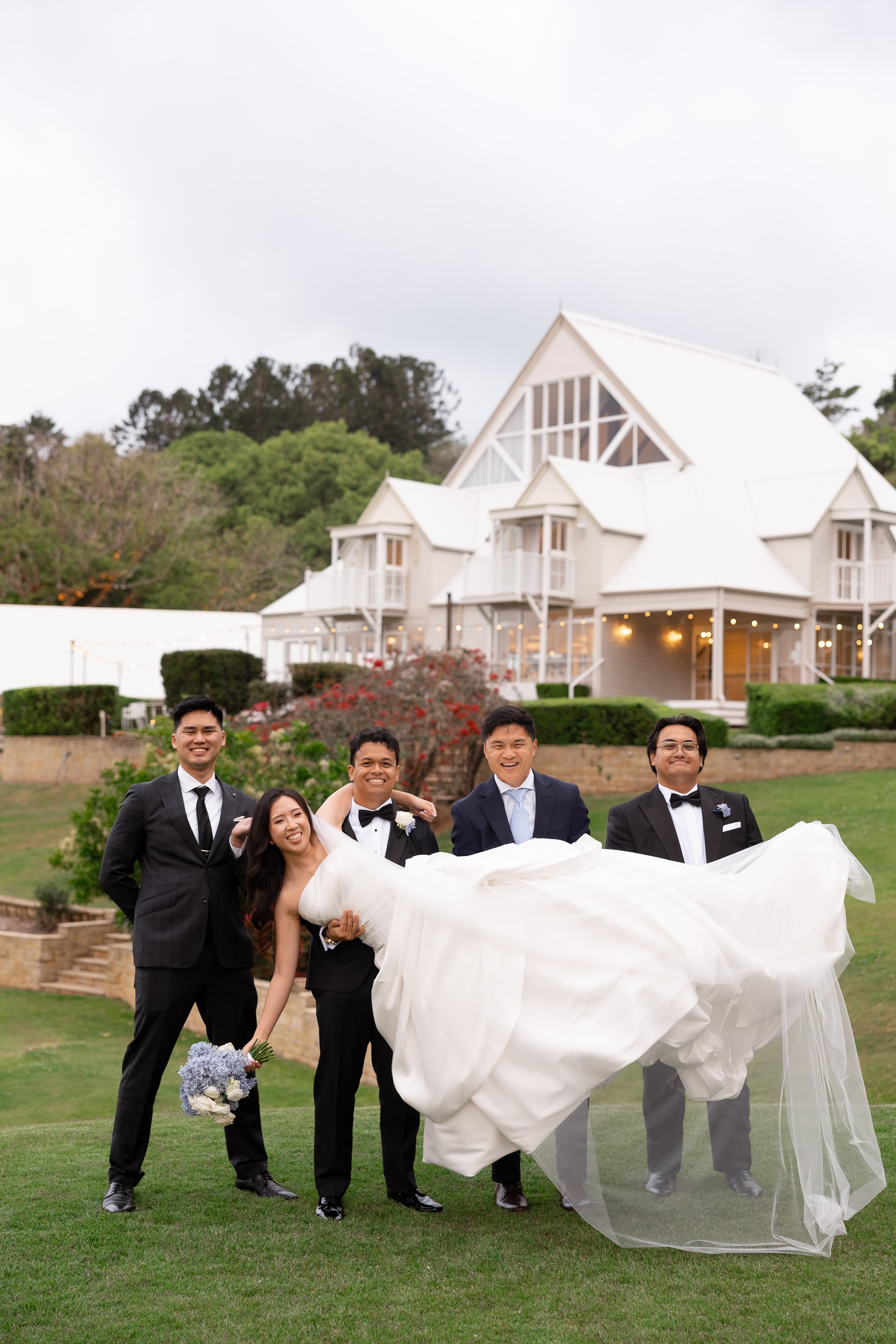 Alicia in her gown carried by Ezra and groomsmen on the lawn in front of Maleny Manor
