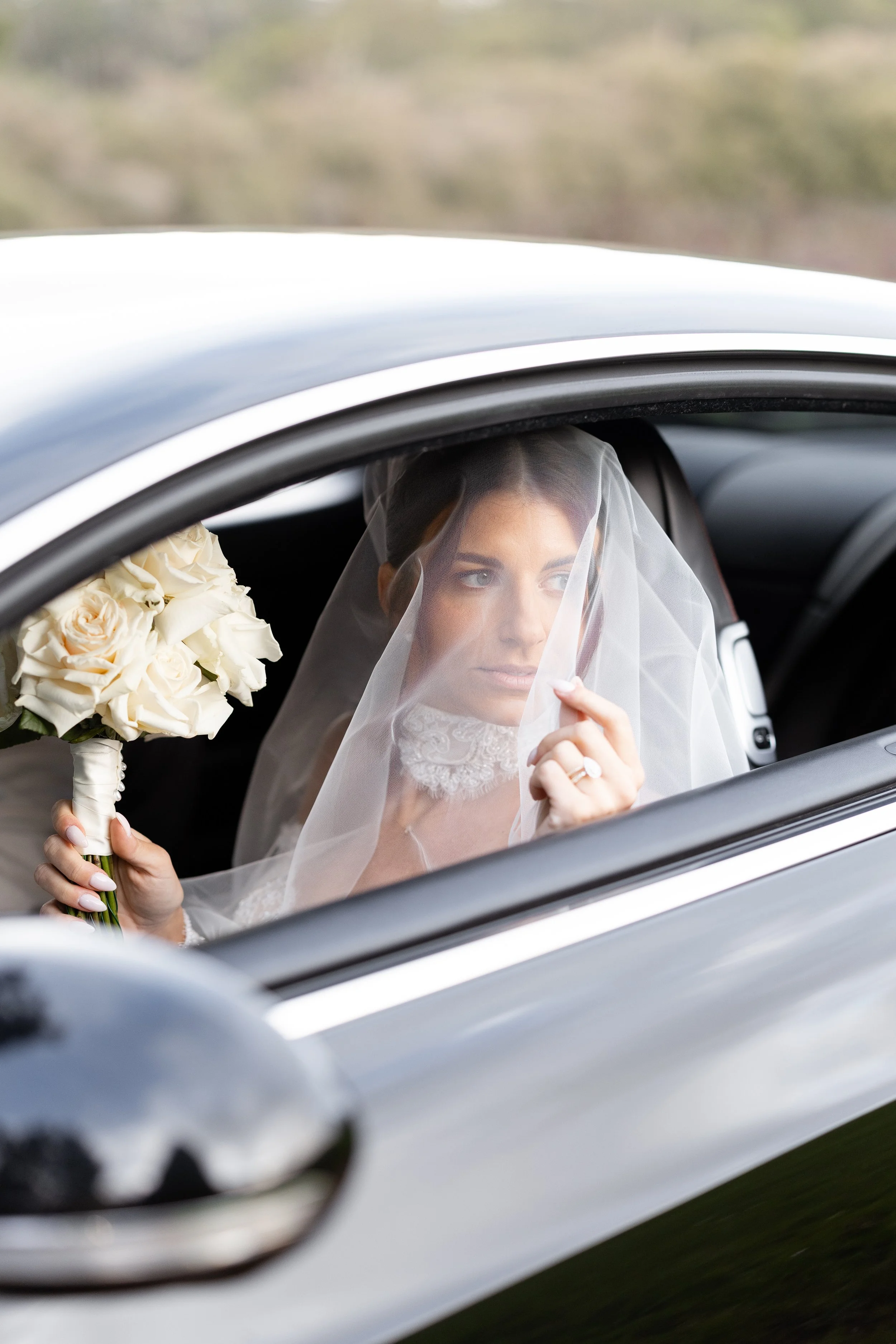 Steph in her veil holding a white bouquet in the back of a car, framed by the window